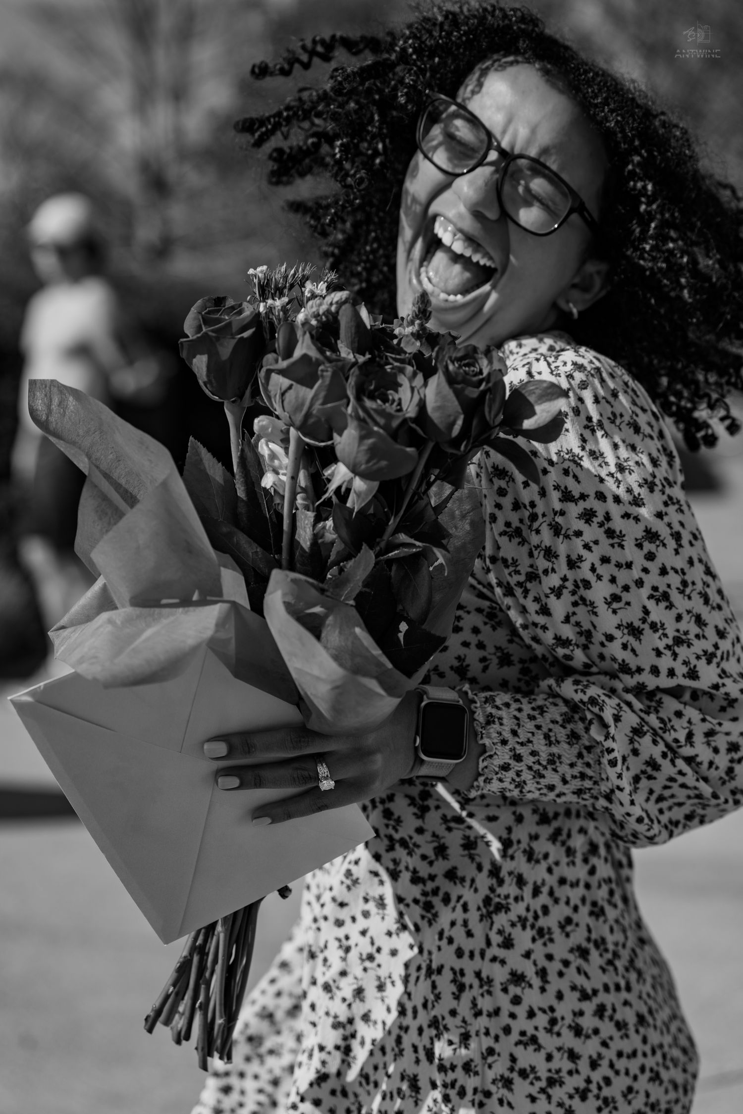 A joyful woman with curly hair and glasses laughs while holding a bouquet of roses in black and white.