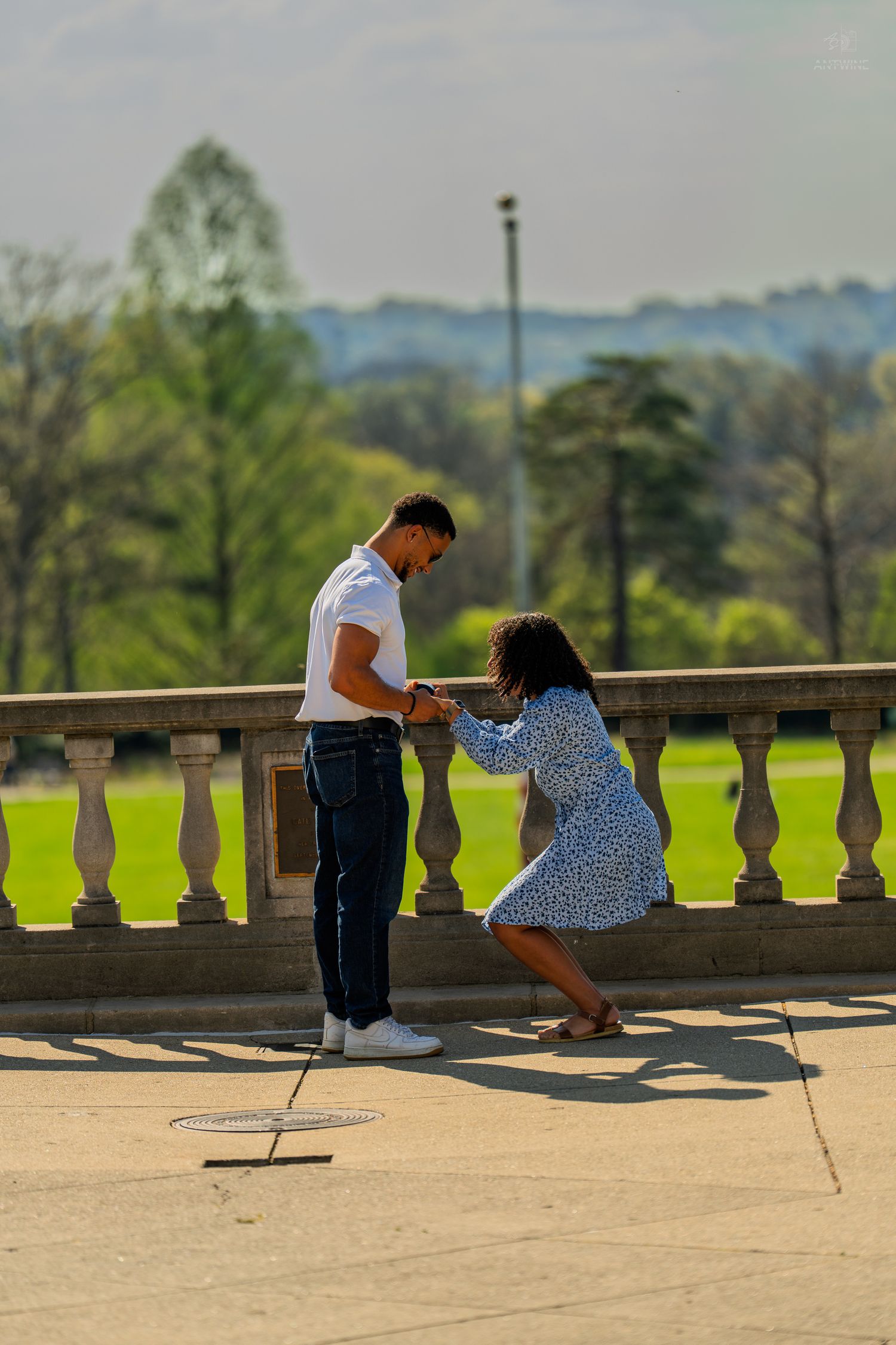 Man and woman share a romantic moment on a stone terrace overlooking a lush green park.