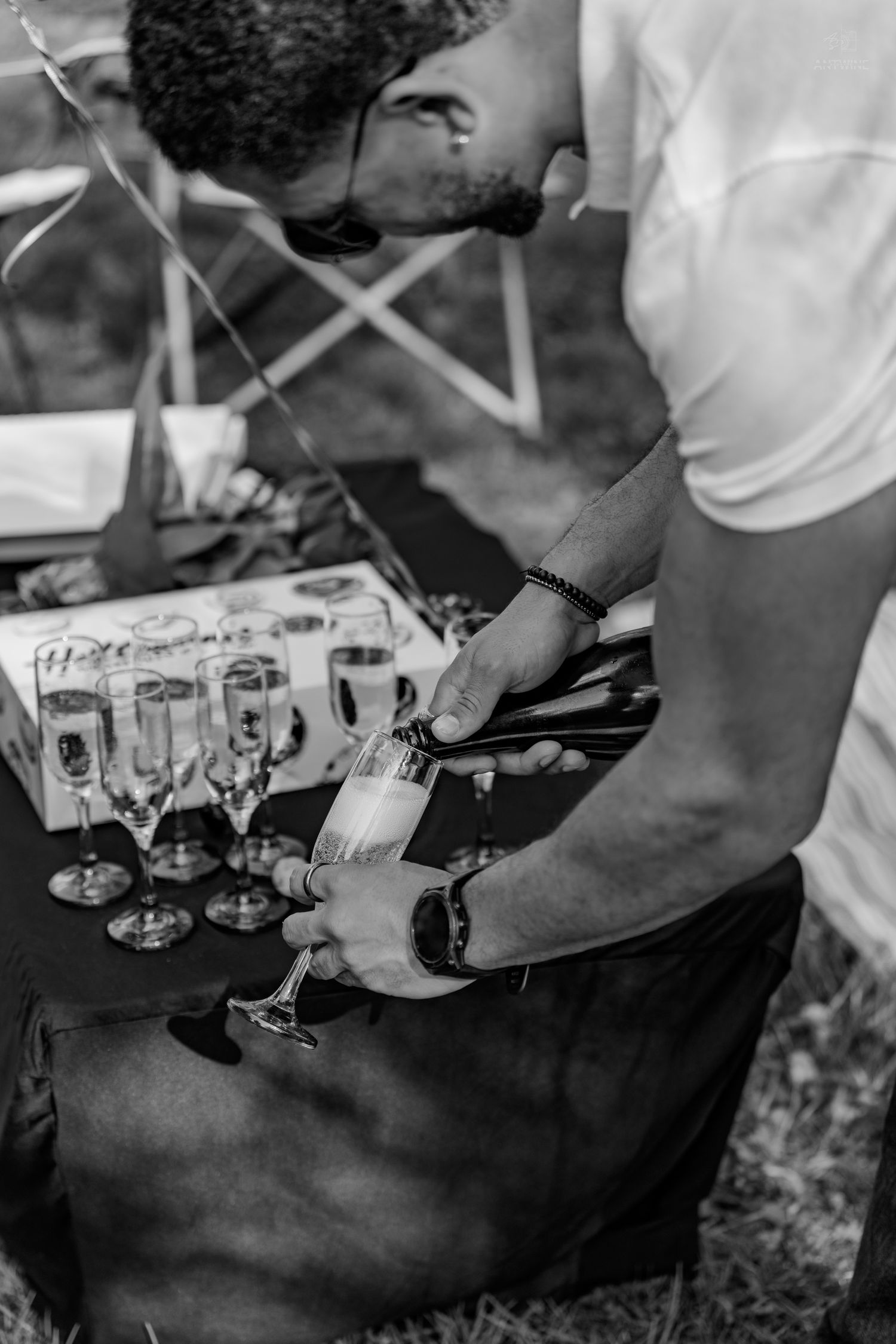 Man carefully pouring champagne into a flute at an outdoor celebration in black and white.
