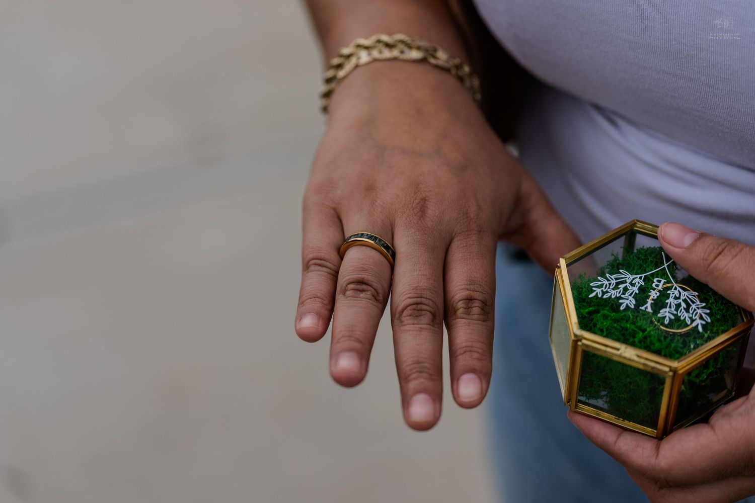 Close-up of a hand with a gold band ring beside a hexagonal glass ring box with moss.