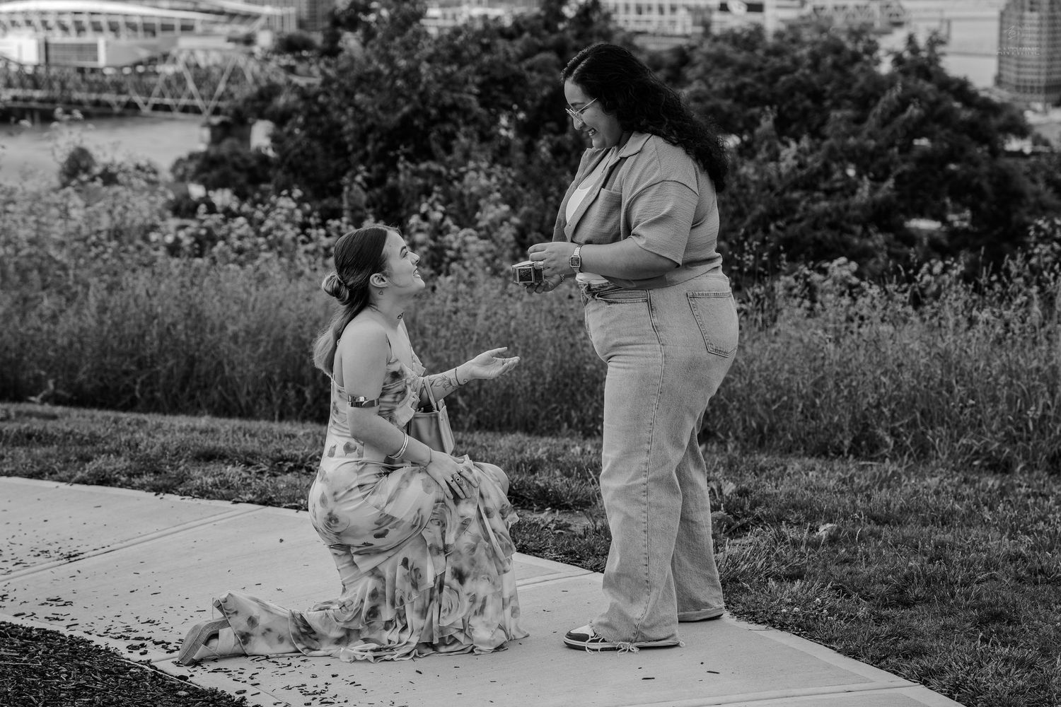 Black and white photo capturing intimate same-sex proposal in park setting.