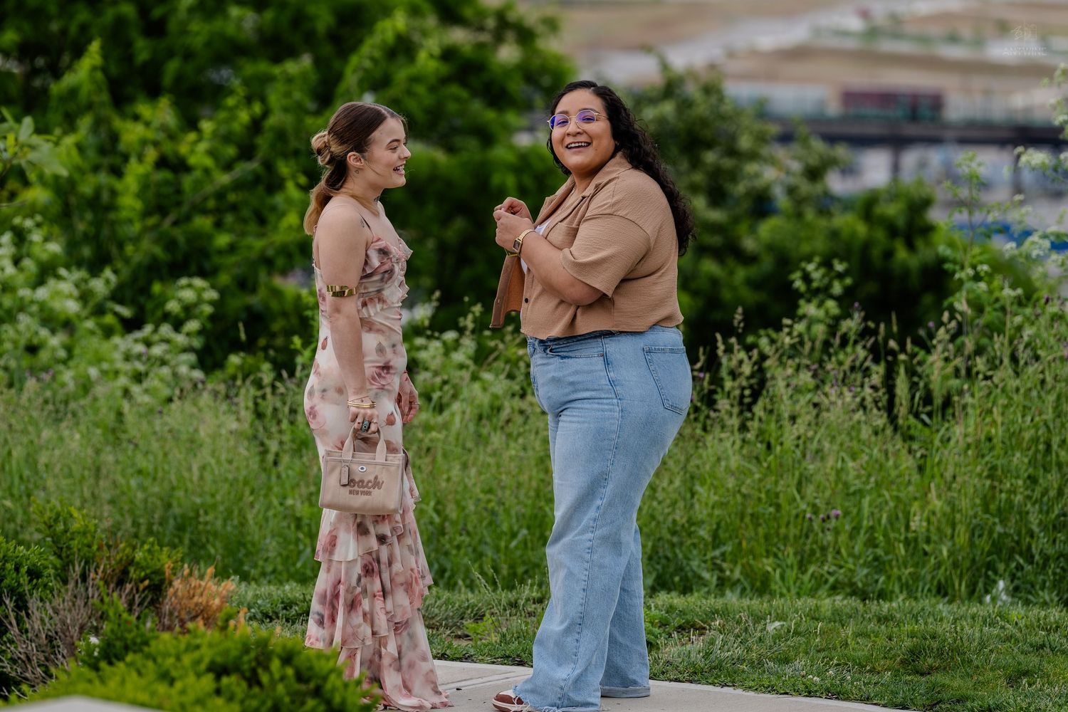 Two women smile and chat outdoors in a garden, one wearing a floral maxi dress and one in a tan top.