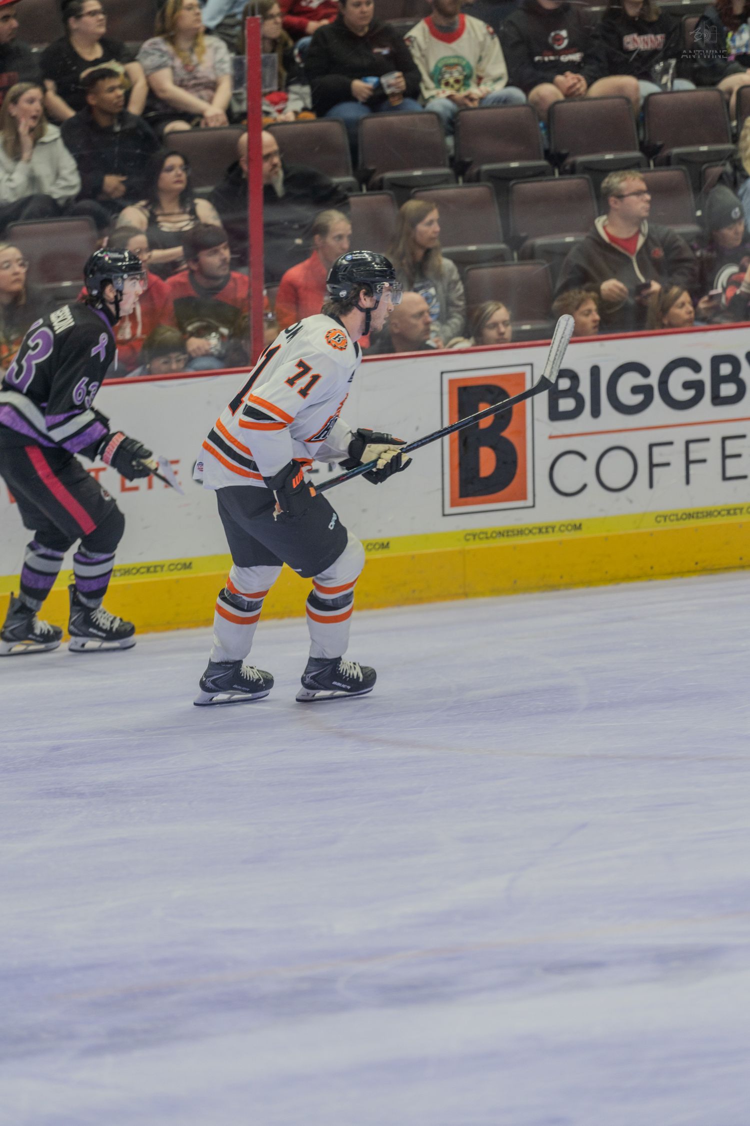 Hockey player #71 in orange and white uniform skates near boards past Biggby Coffee advertisement during live game.