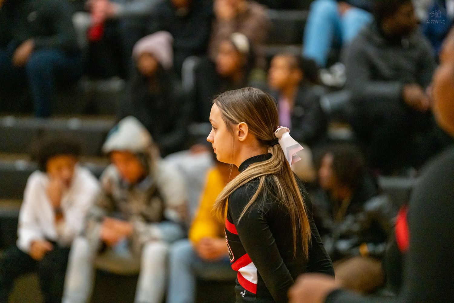 A cheerleader in a black uniform stands attentively on the sideline during a high school basketball game.