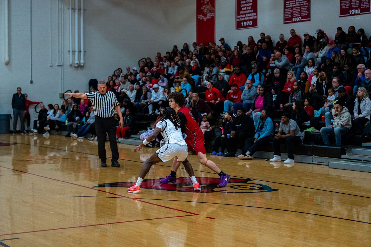 Two basketball players battle for possession on the court during a high school game with a packed crowd.