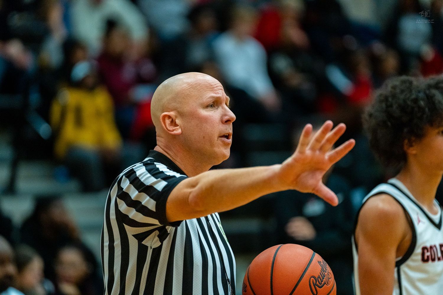 Bald basketball referee raises hand to signal call during indoor game.