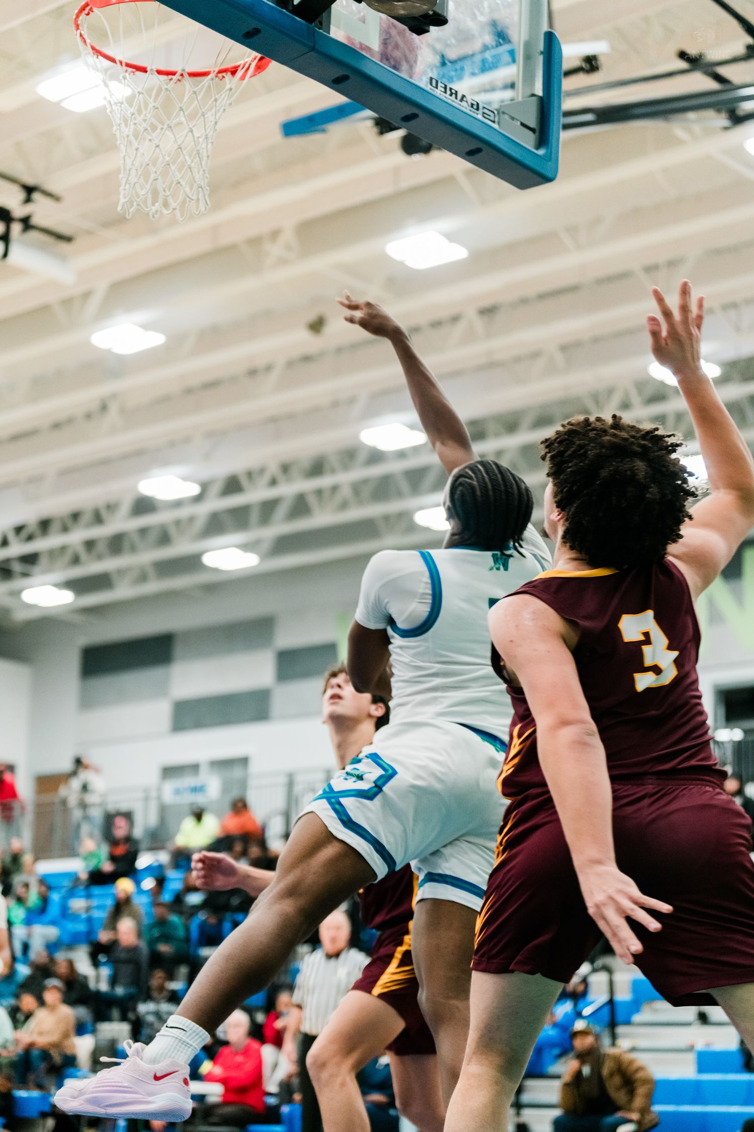 A white-uniformed player drives to the basket past maroon #3 defender during a high school basketball game.