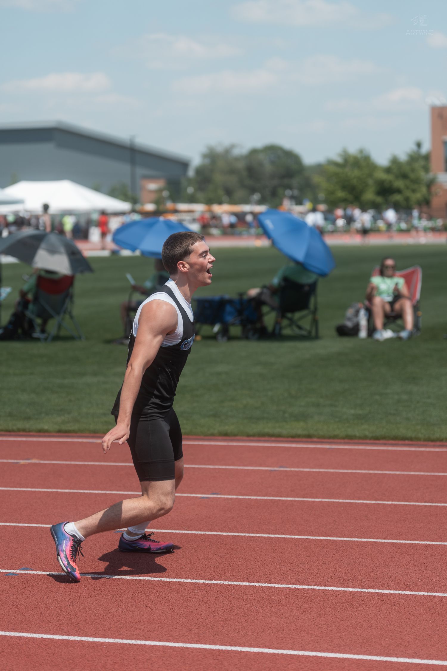Smiling male athlete in black uniform walks on a red track at an outdoor track and field competition on a sunny day.