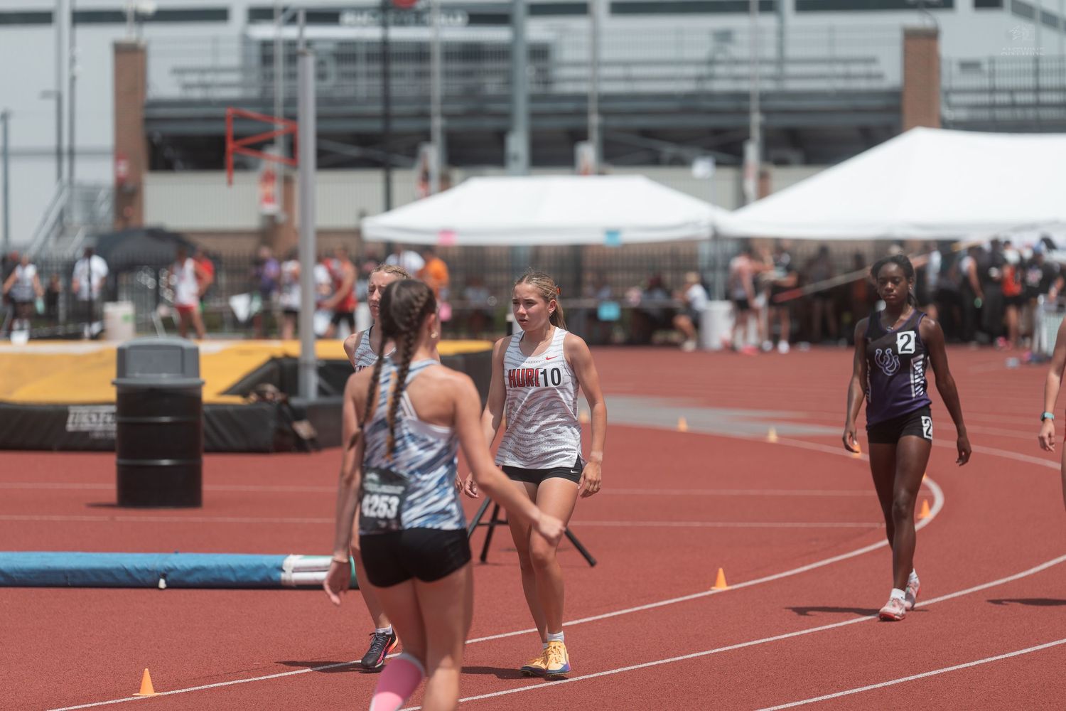 Female relay runners exchanging baton on red track during sunny outdoor high school track and field competition.