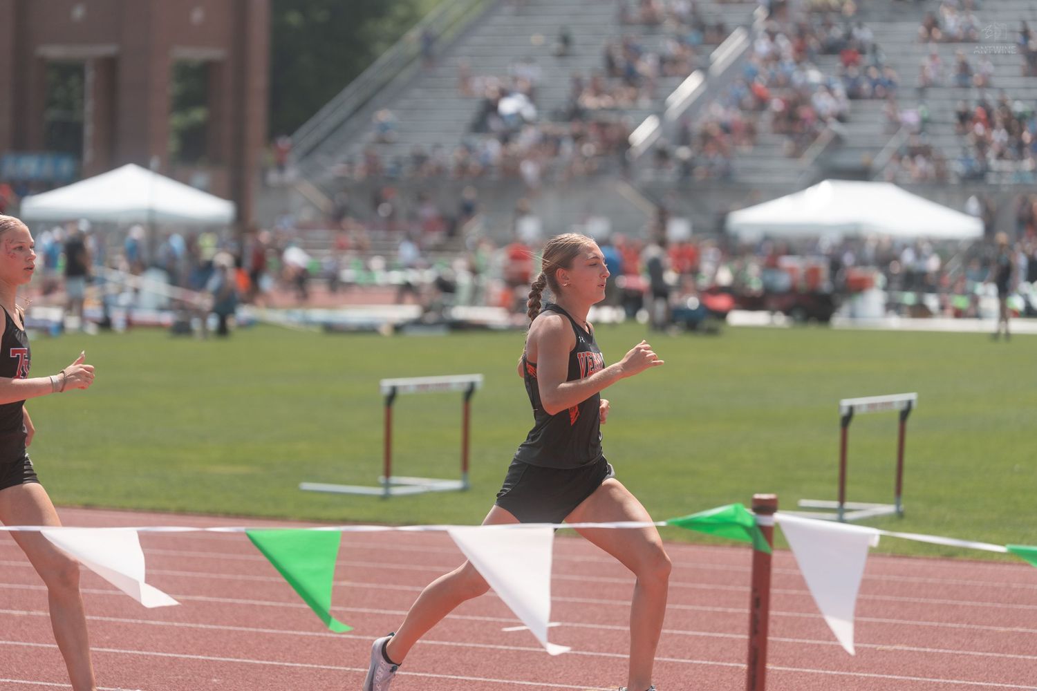 A female runner in black competes past green flags at an outdoor track and field event.