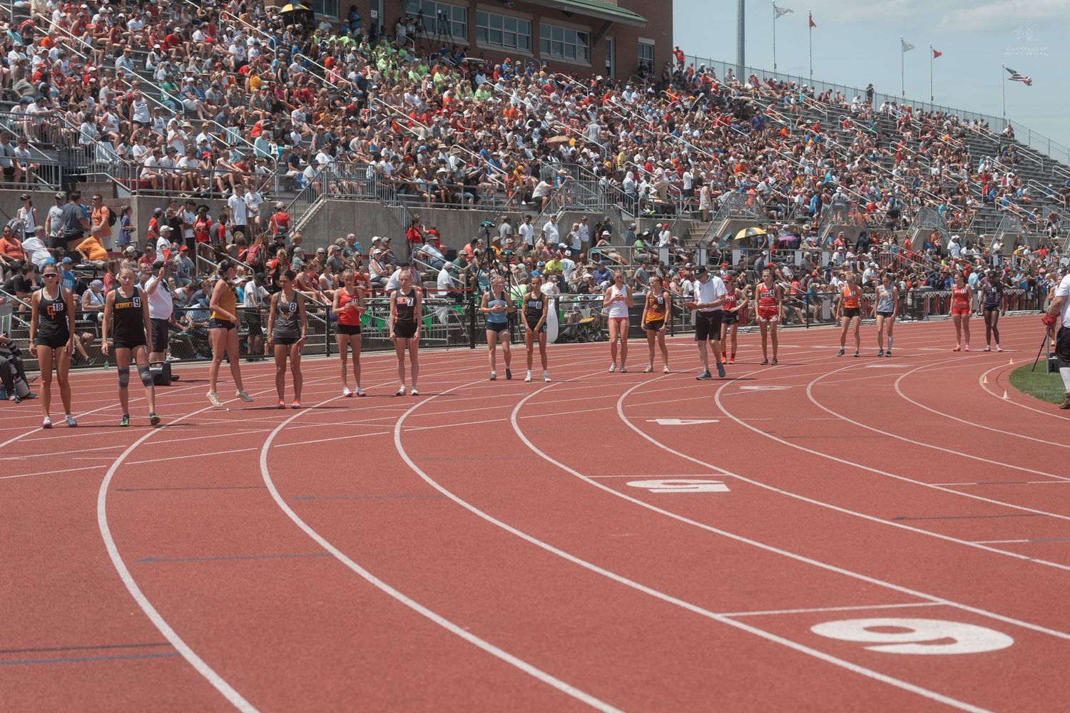 Female athletes line up at the start of a track race in a packed stadium during a high school track meet.