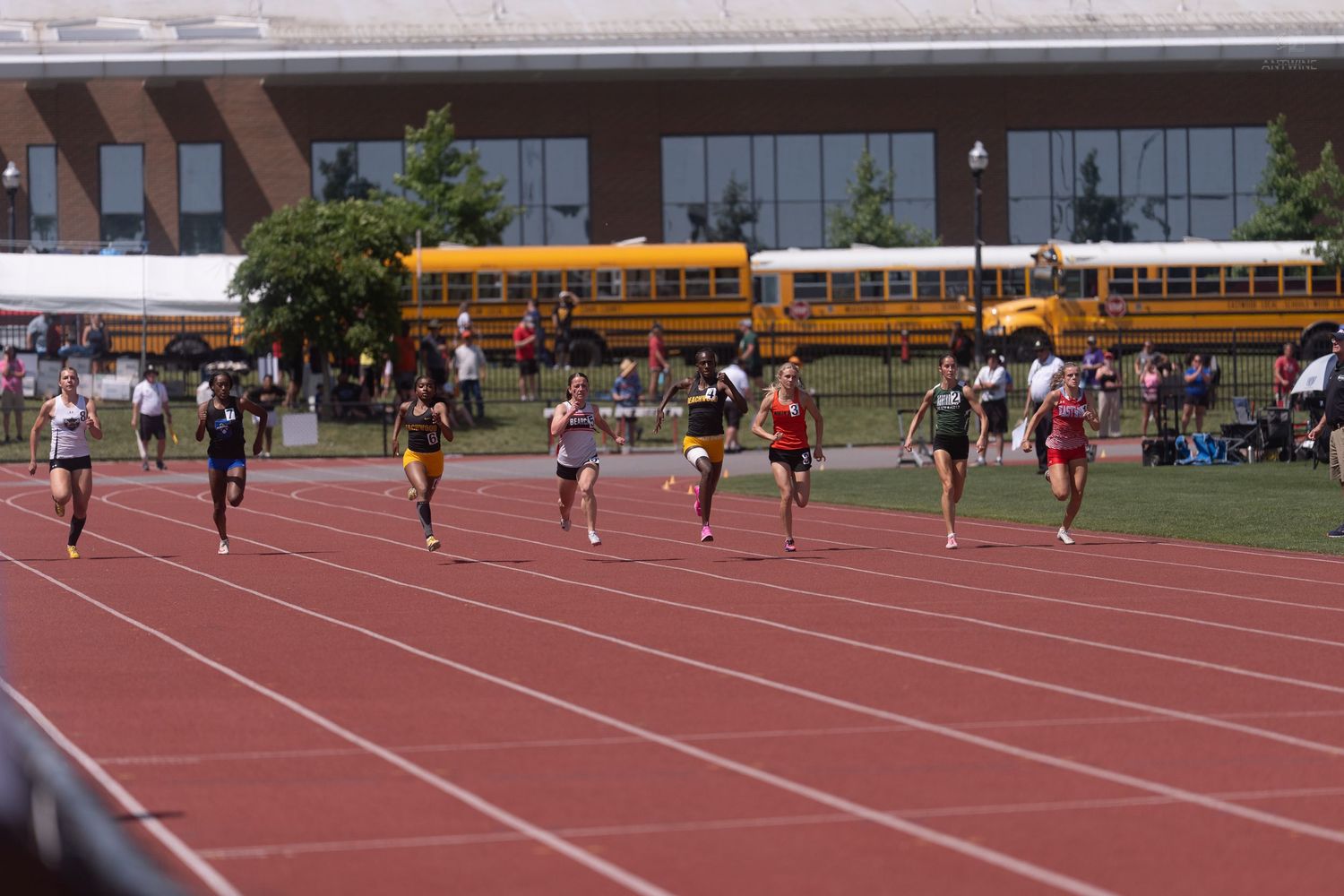 Female sprinters race down the track at an outdoor meet with school buses visible in the background.