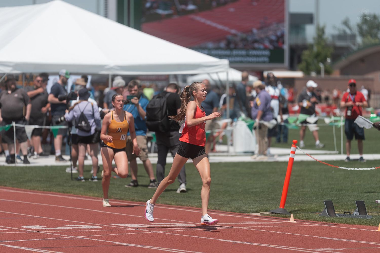 Female runners compete on a red track during an outdoor track and field competition on a sunny day.
