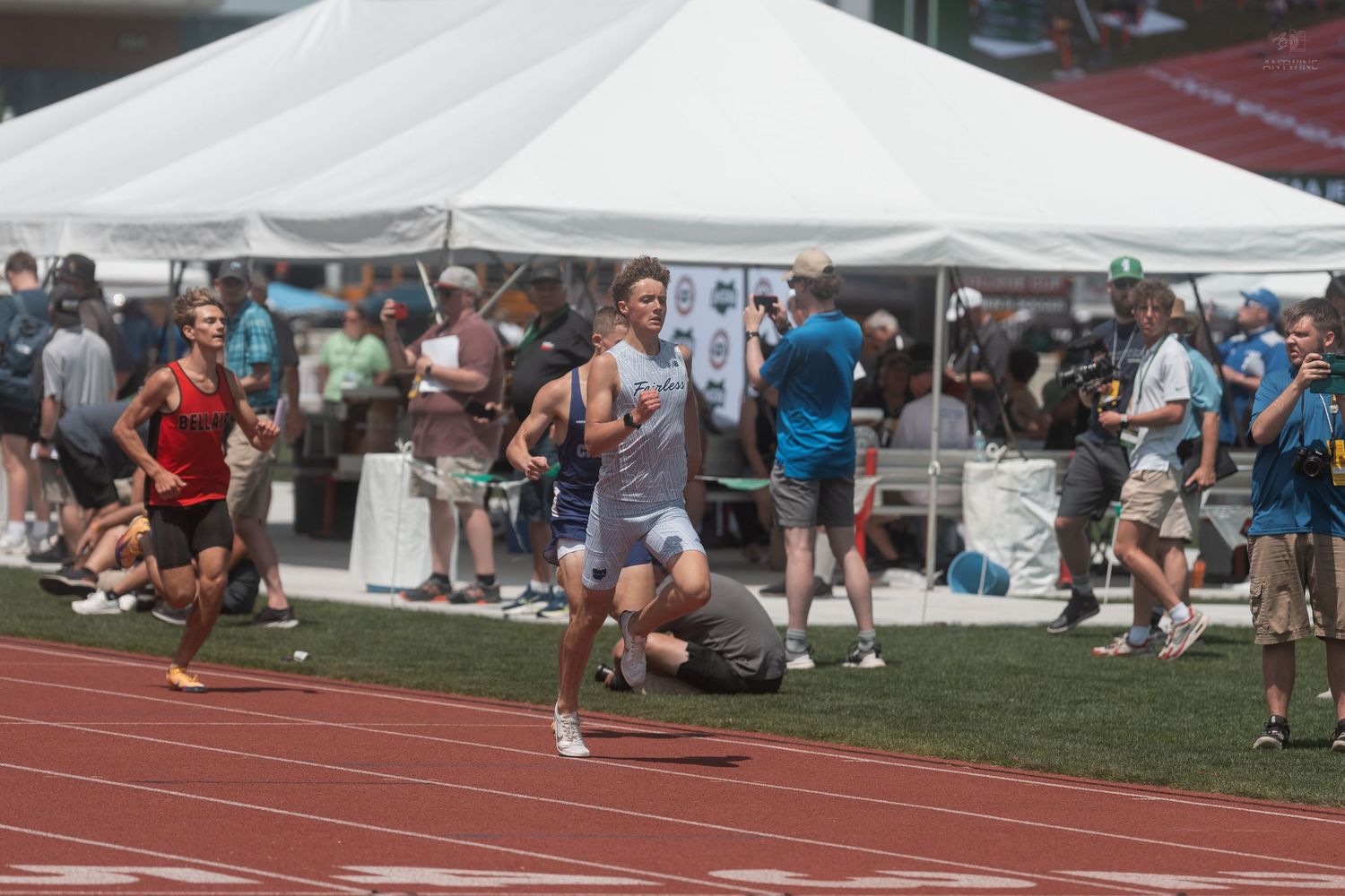 Athletes compete in a middle distance track race at an outdoor stadium during a sunny day championship meet.