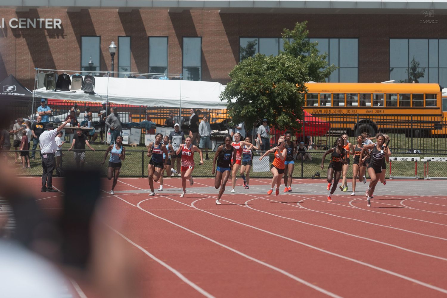 Female athletes sprinting on red track at outdoor collegiate track and field championship near brick building.