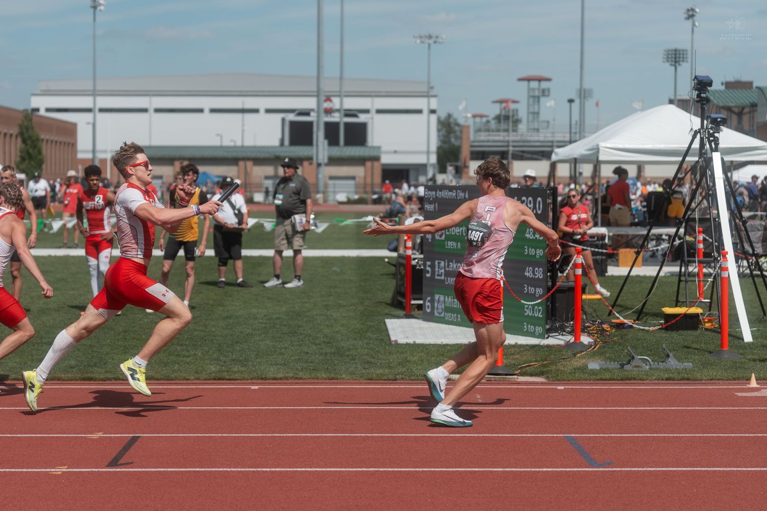 Athletes exchange baton during relay race on red track at outdoor high school track and field competition.