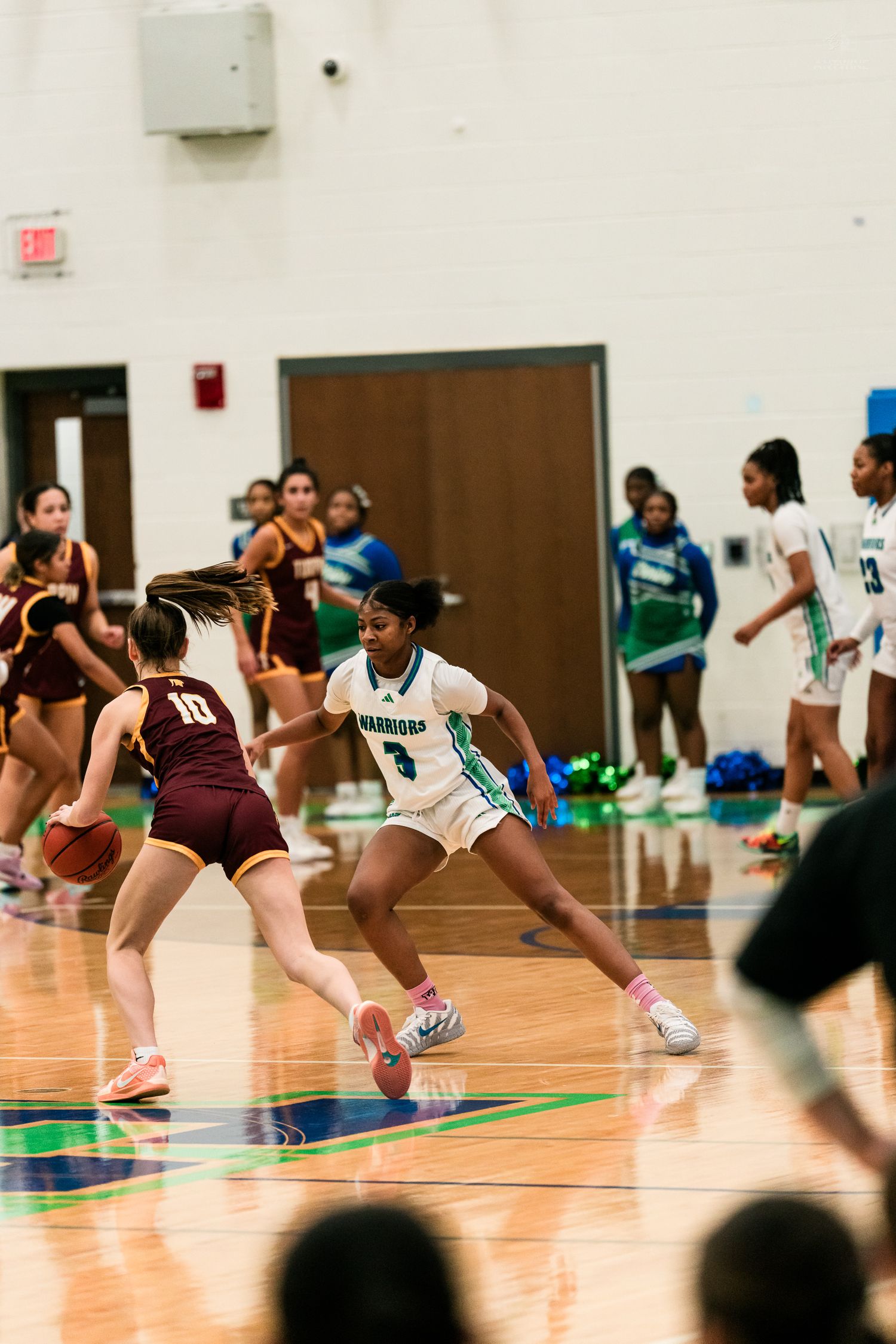 A Warriors player number 3 in teal plays defense against a maroon number 10 opponent in girls basketball.