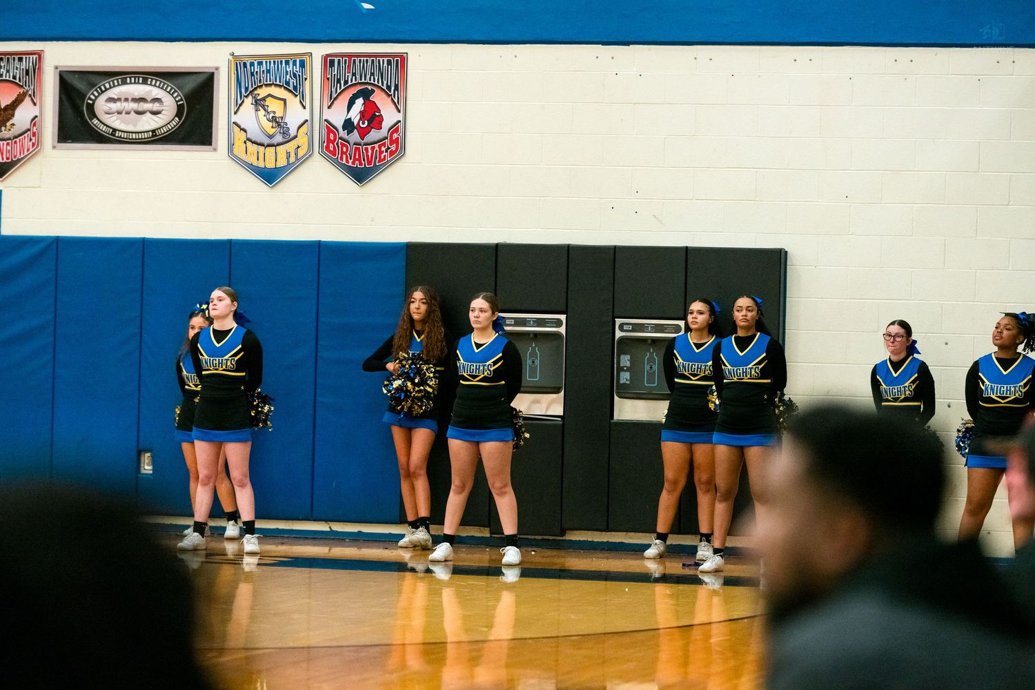 High school cheerleaders in blue and black uniforms stand in a line along the gymnasium wall during a basketball game.