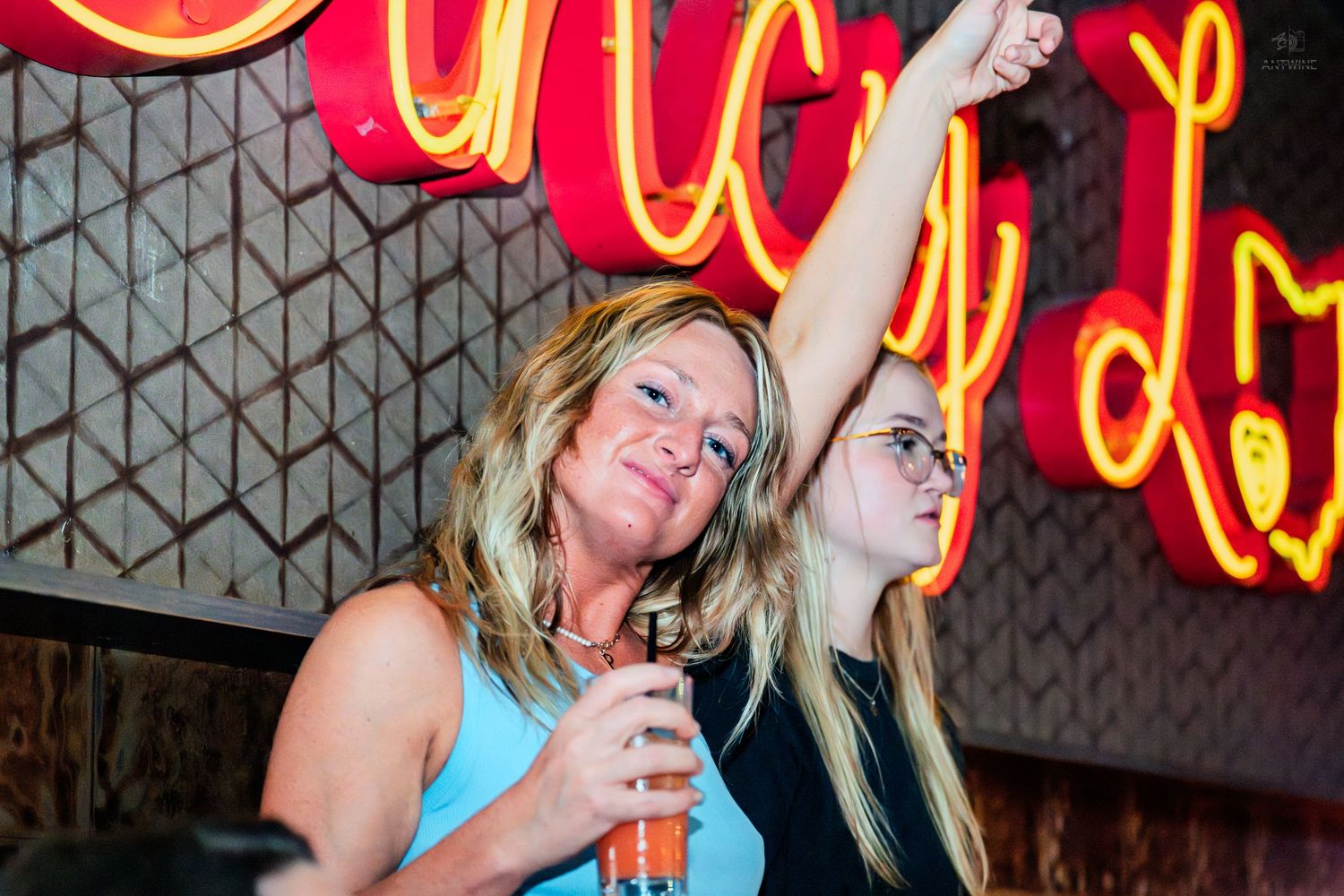Two women celebrating at a bar with neon signs, one raising her arm holding a drink.