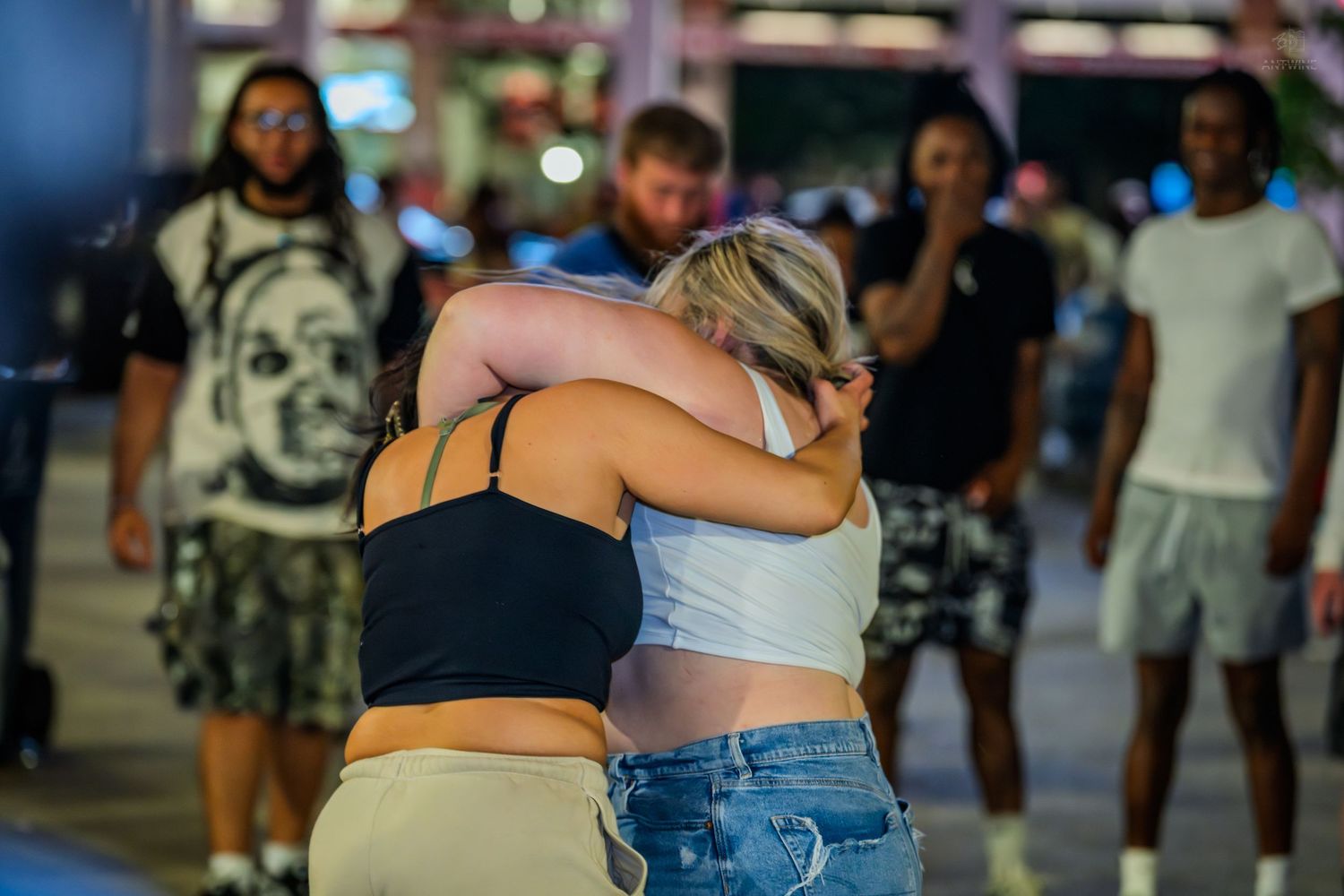 Two women embracing in a comforting hug on a busy nighttime street outdoors.