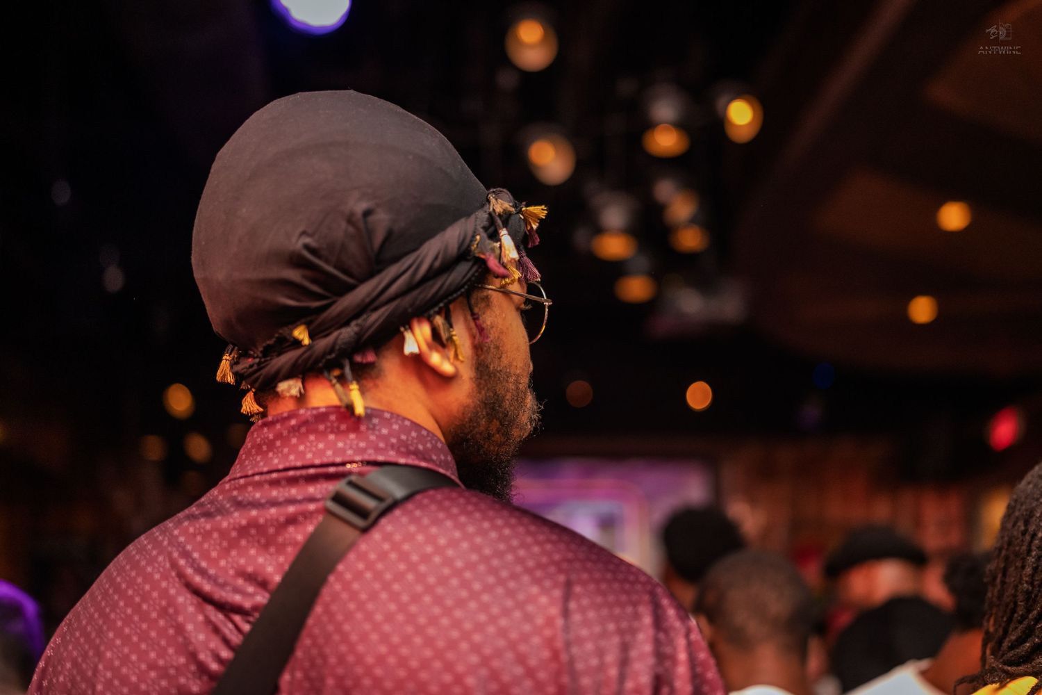 Back view of a man wearing a durag and patterned shirt at a crowded music venue.