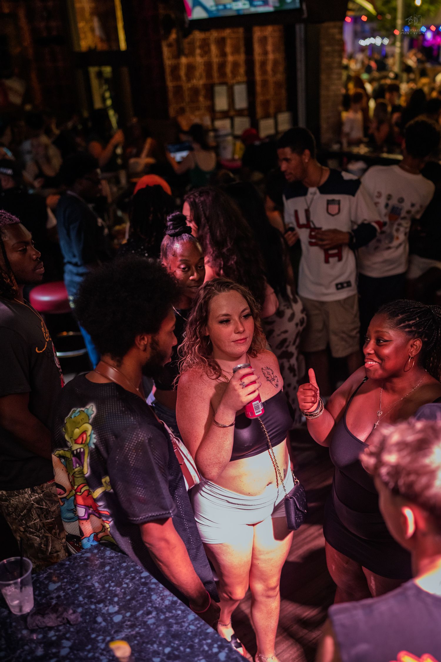 Woman in crop top holding drink socializes with friends at a crowded nightclub bar event.