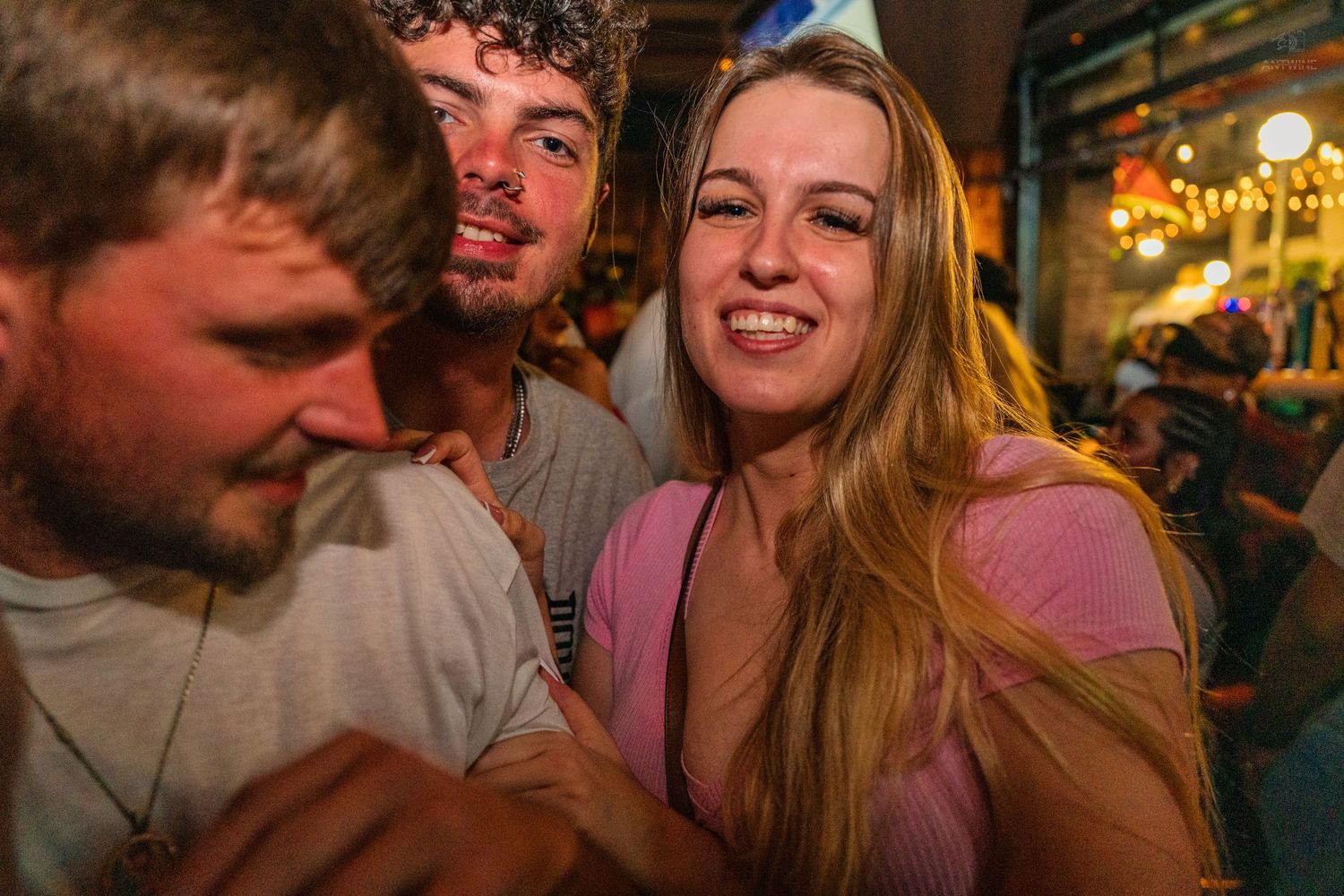 Three young people smiling and posing closely together at a lively outdoor nightlife event.