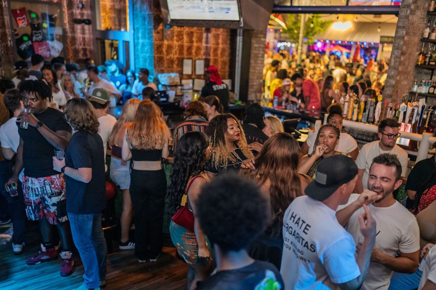 Busy bar crowd with people mingling and dancing near a well-stocked bar with colorful lighting.