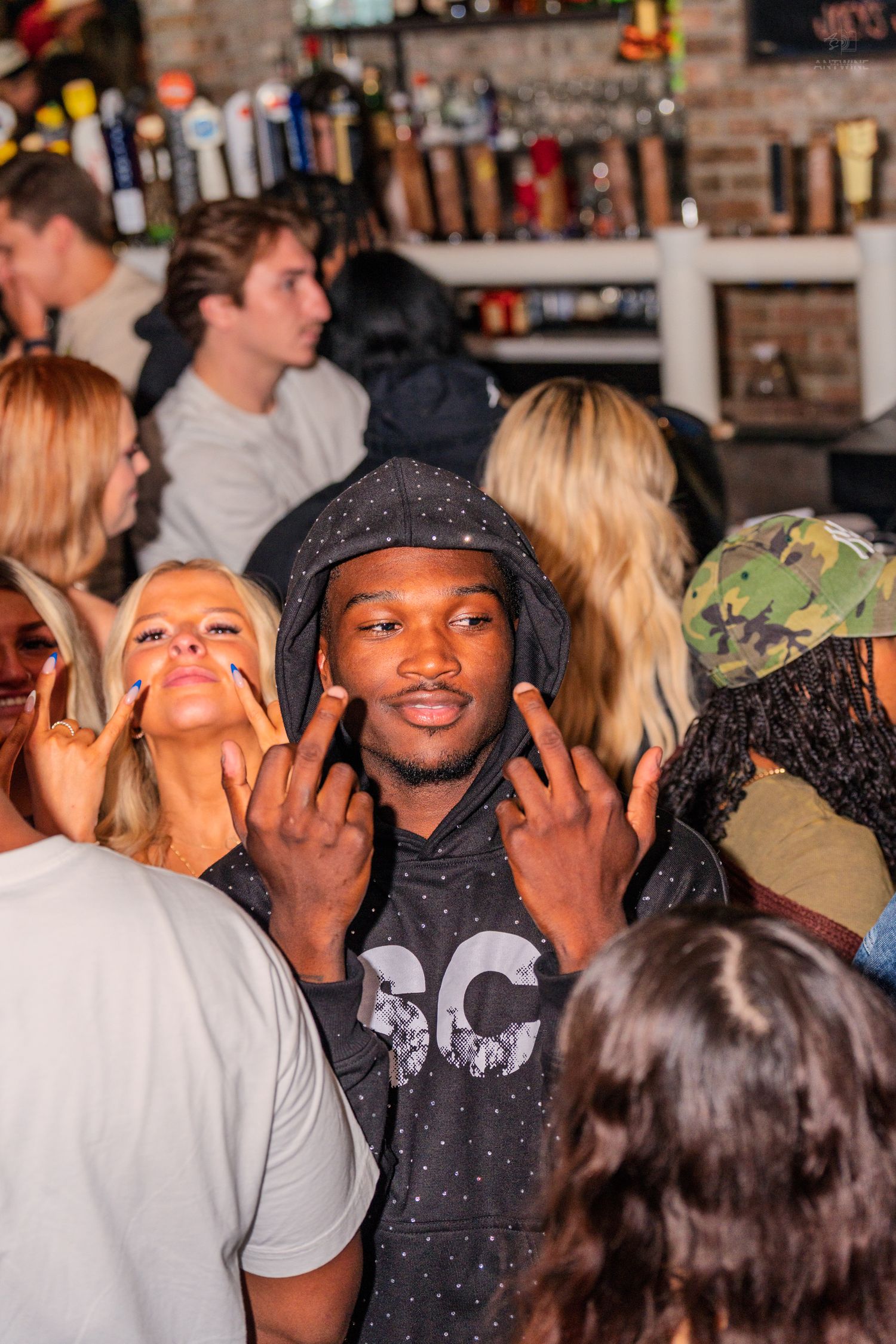 Man in dark hoodie making hand gestures surrounded by crowd at a lively bar event.