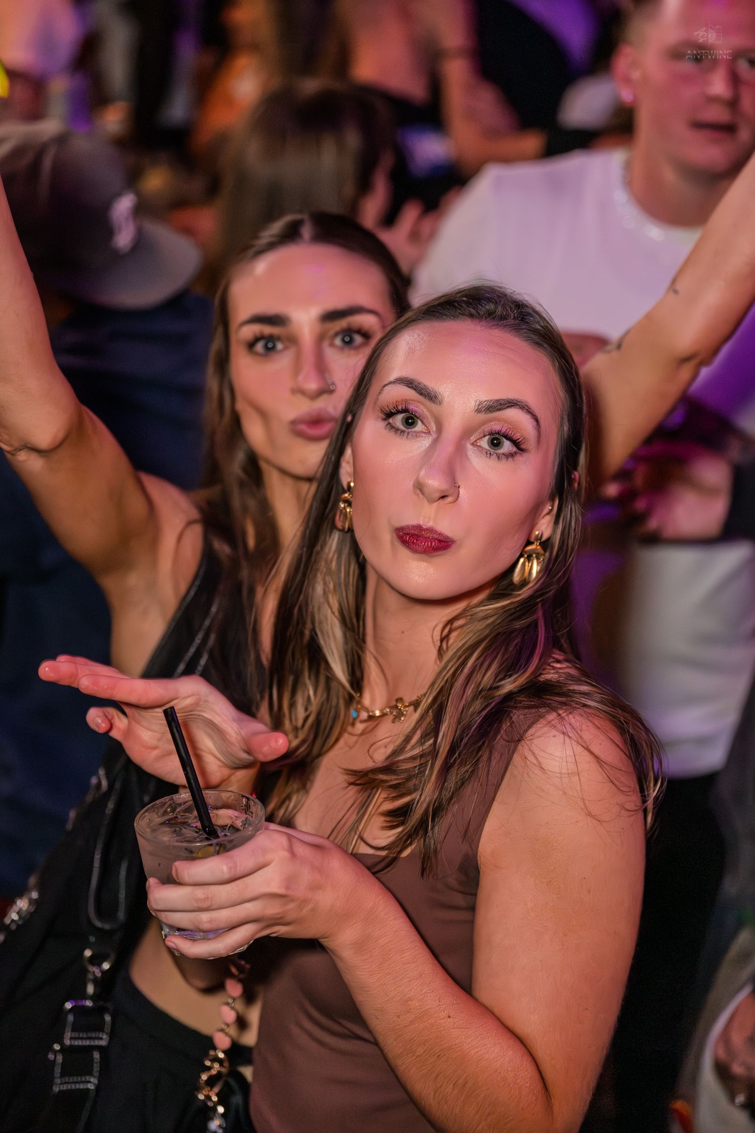 Two young women posing and having fun at a crowded nightclub party with drinks.
