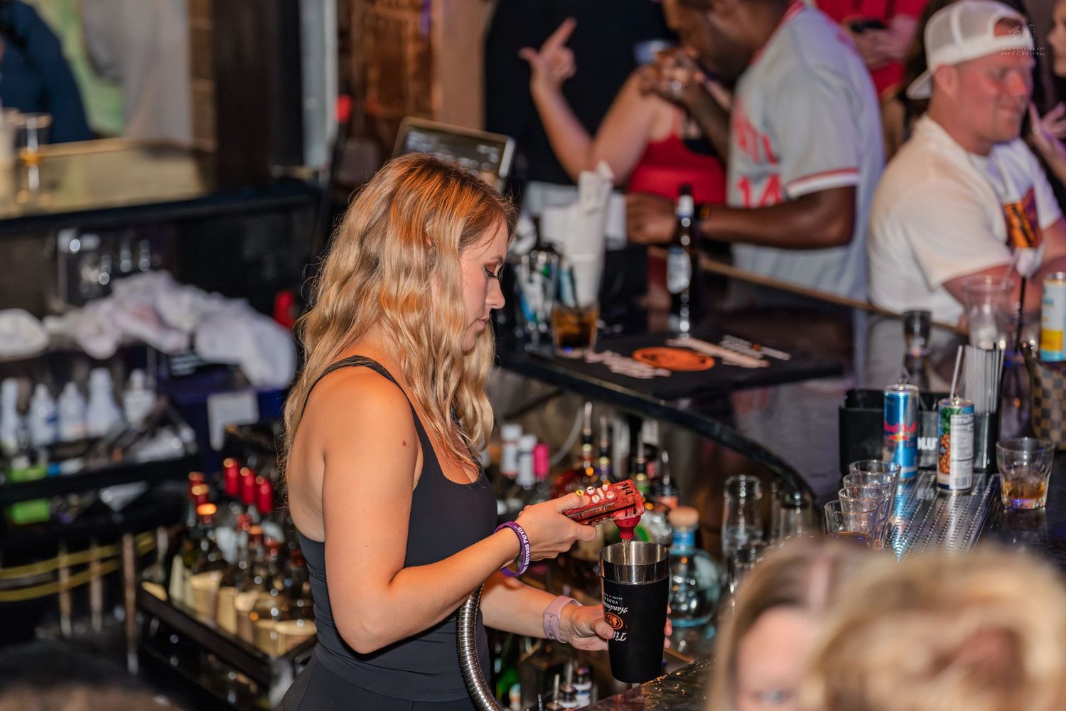 Female bartender in black top checking her phone behind a well-stocked bar during a busy evening shift.