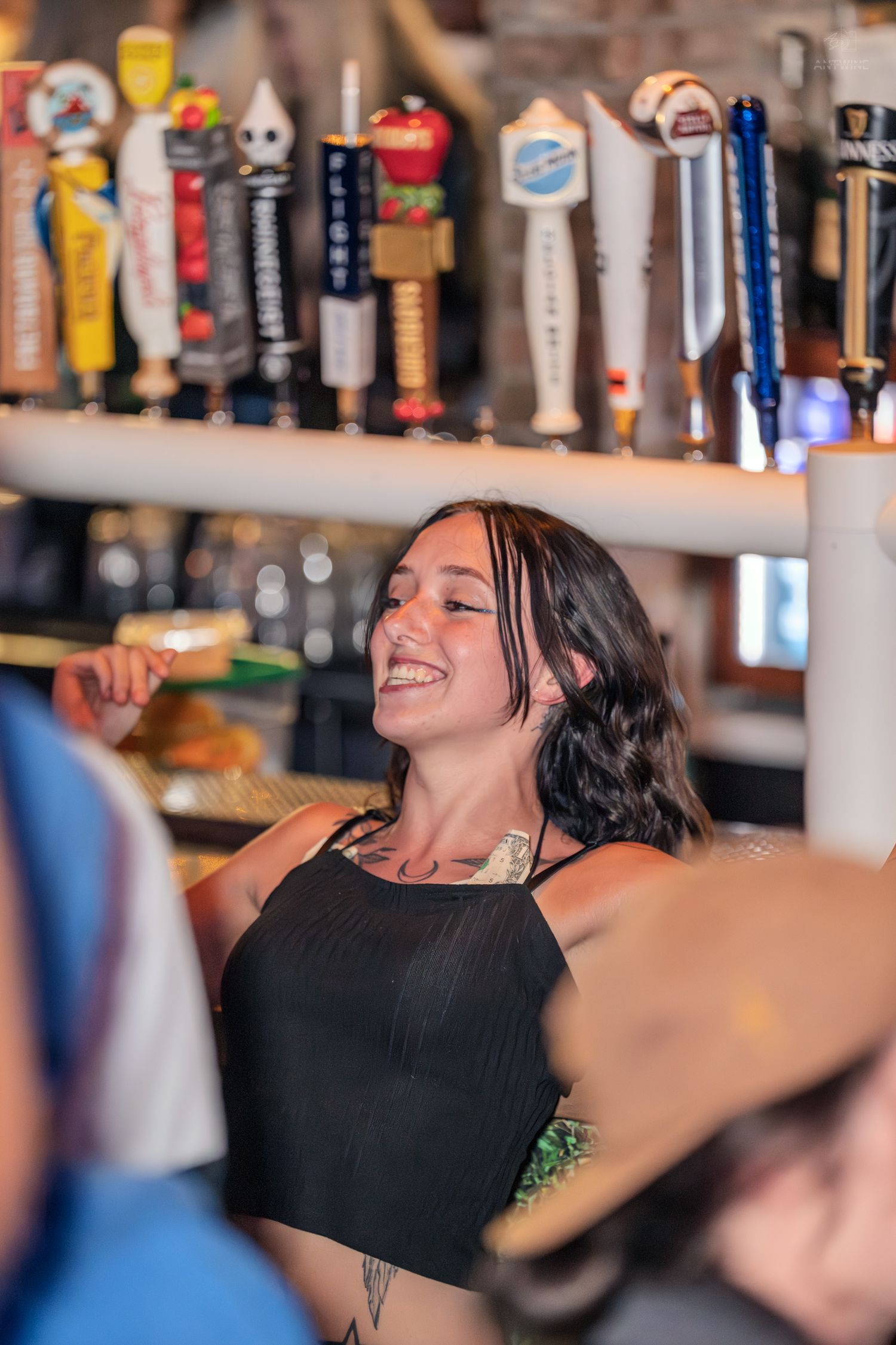 Smiling young woman with dark hair and tattoos standing near beer taps at a busy bar.