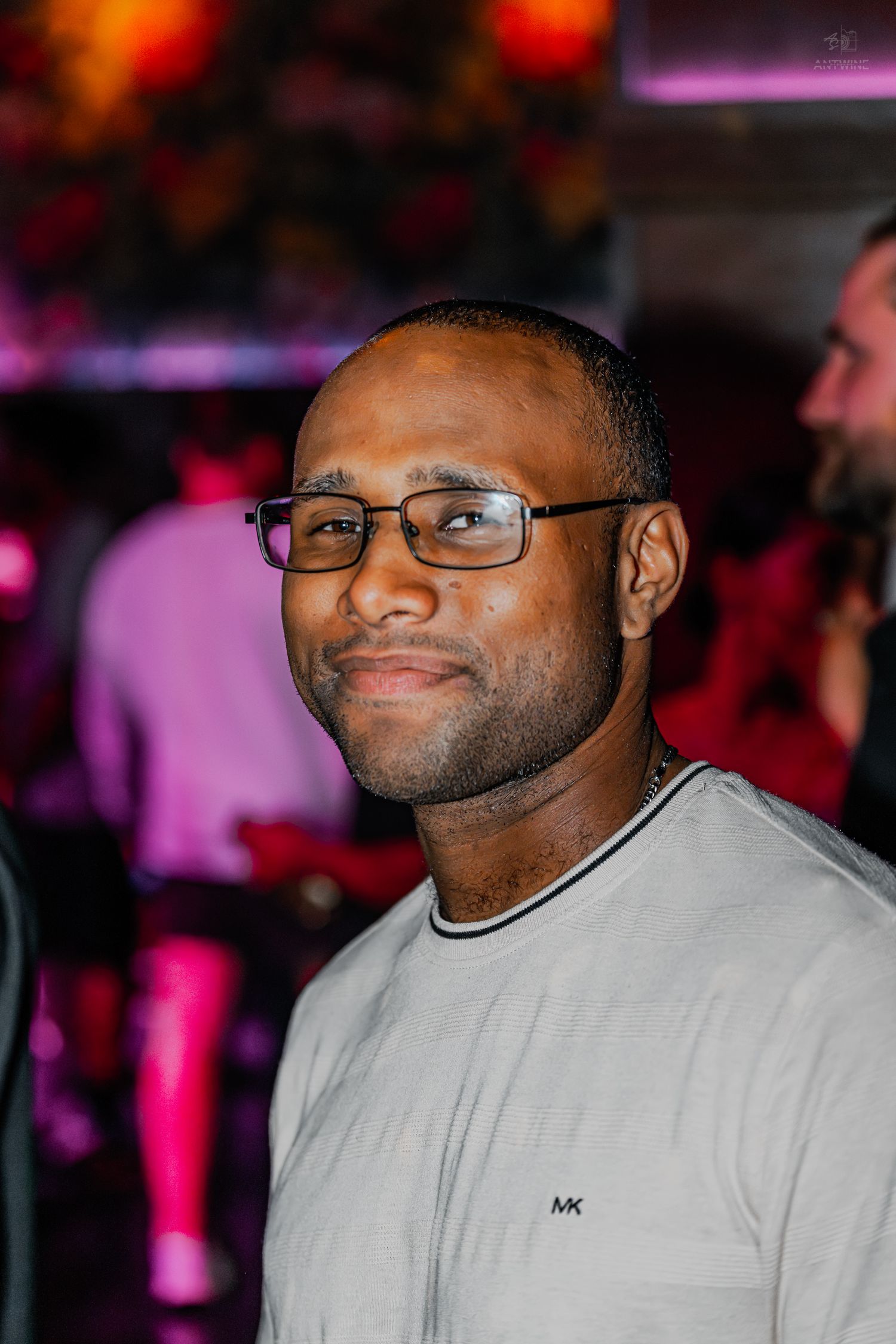 Man wearing glasses and a gray Michael Kors shirt smiling at a vibrant nightclub event.