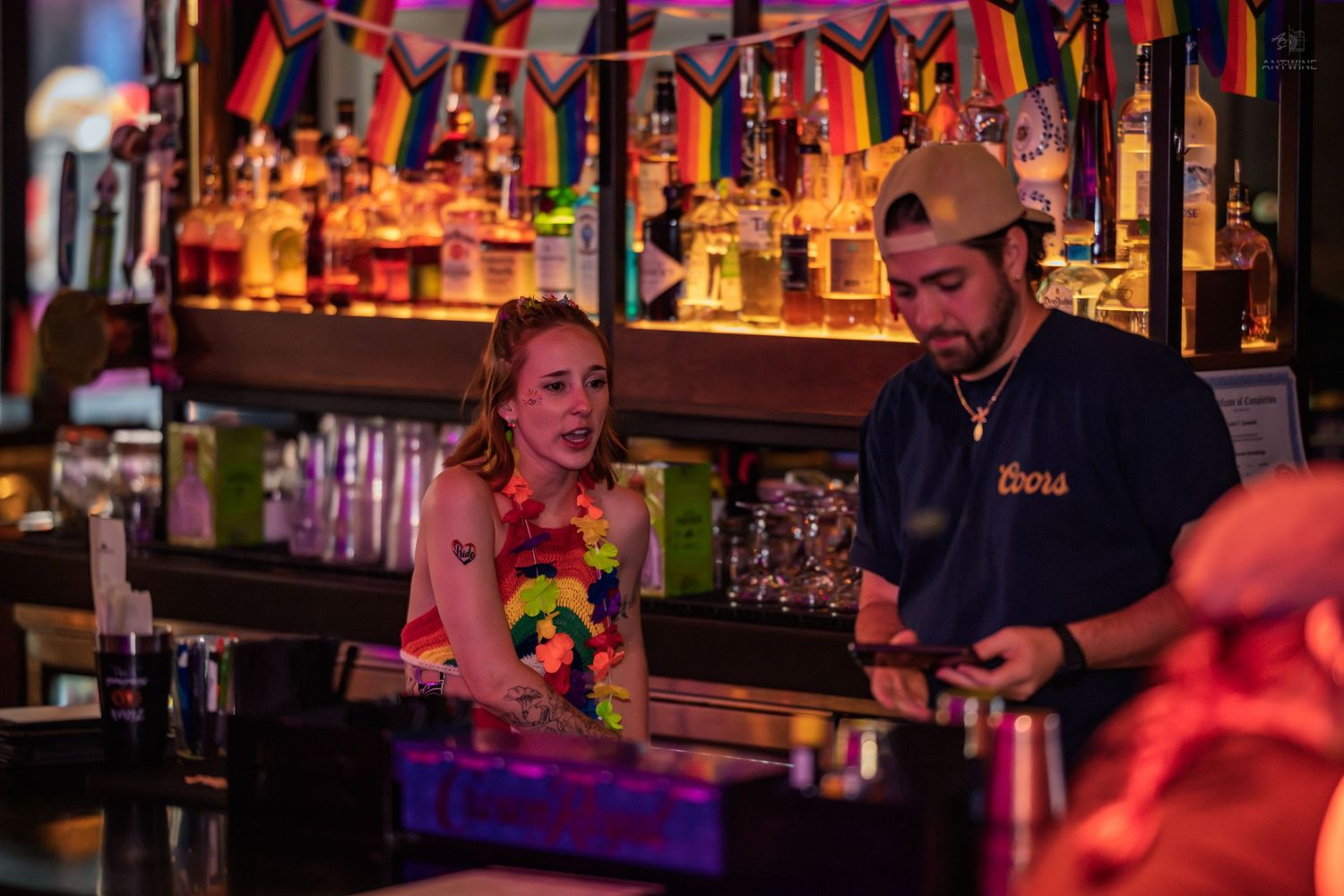 Bartender in cap preparing drinks for a woman at a pride-decorated bar.