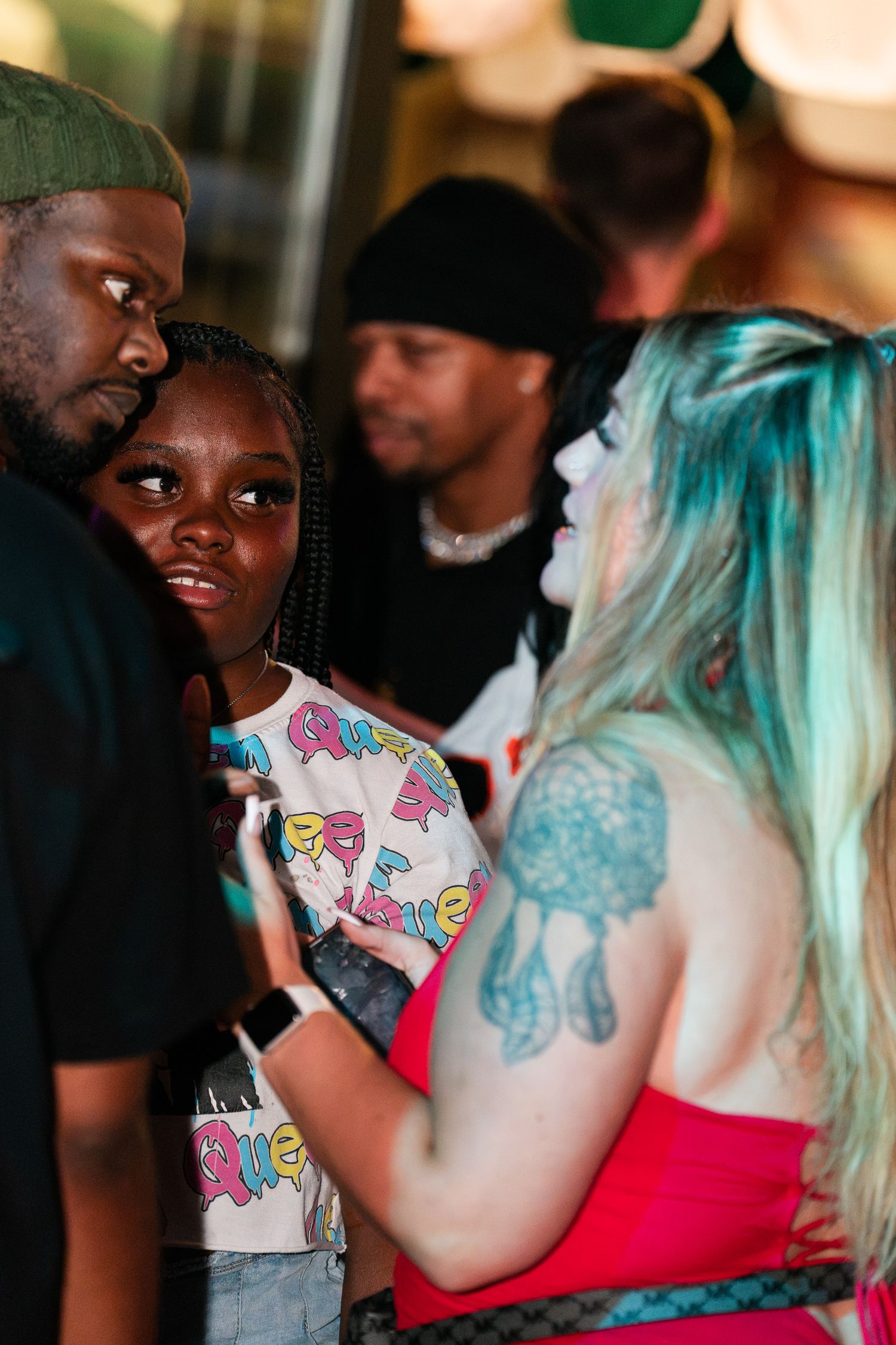 A woman with teal hair and dreamcatcher tattoo talks to a young girl and man at a colorful nightclub event.