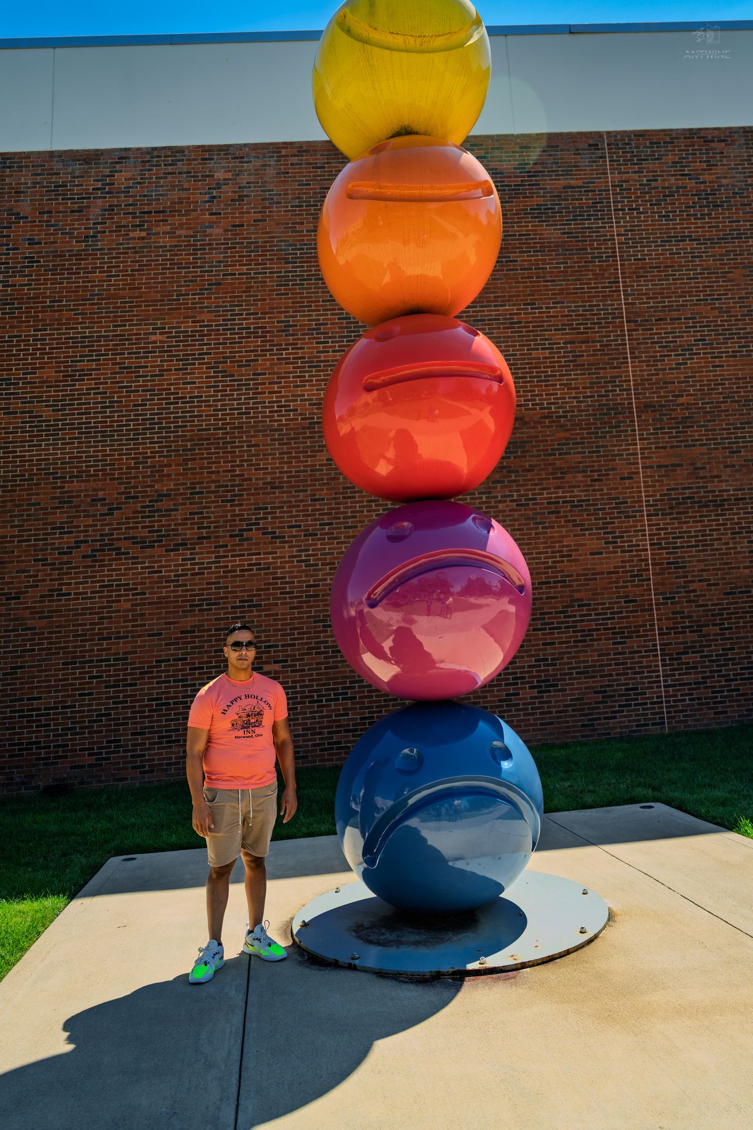 A man in an orange shirt stands beside a tall colorful stacked spheres sculpture against a brick wall.