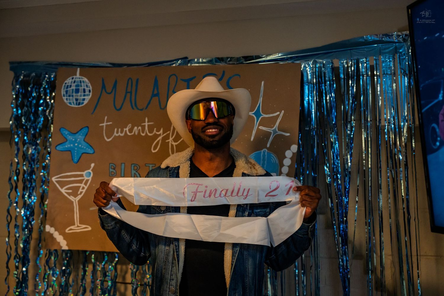 Man in cowboy hat and rainbow sunglasses holding a 'Finally 21' sash at a birthday party.