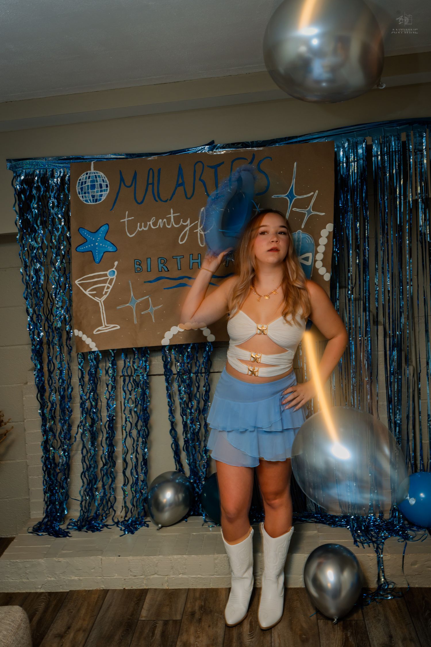 Birthday girl in blue cowboy hat, white boots, and blue skirt poses with silver balloons at 21st birthday party.