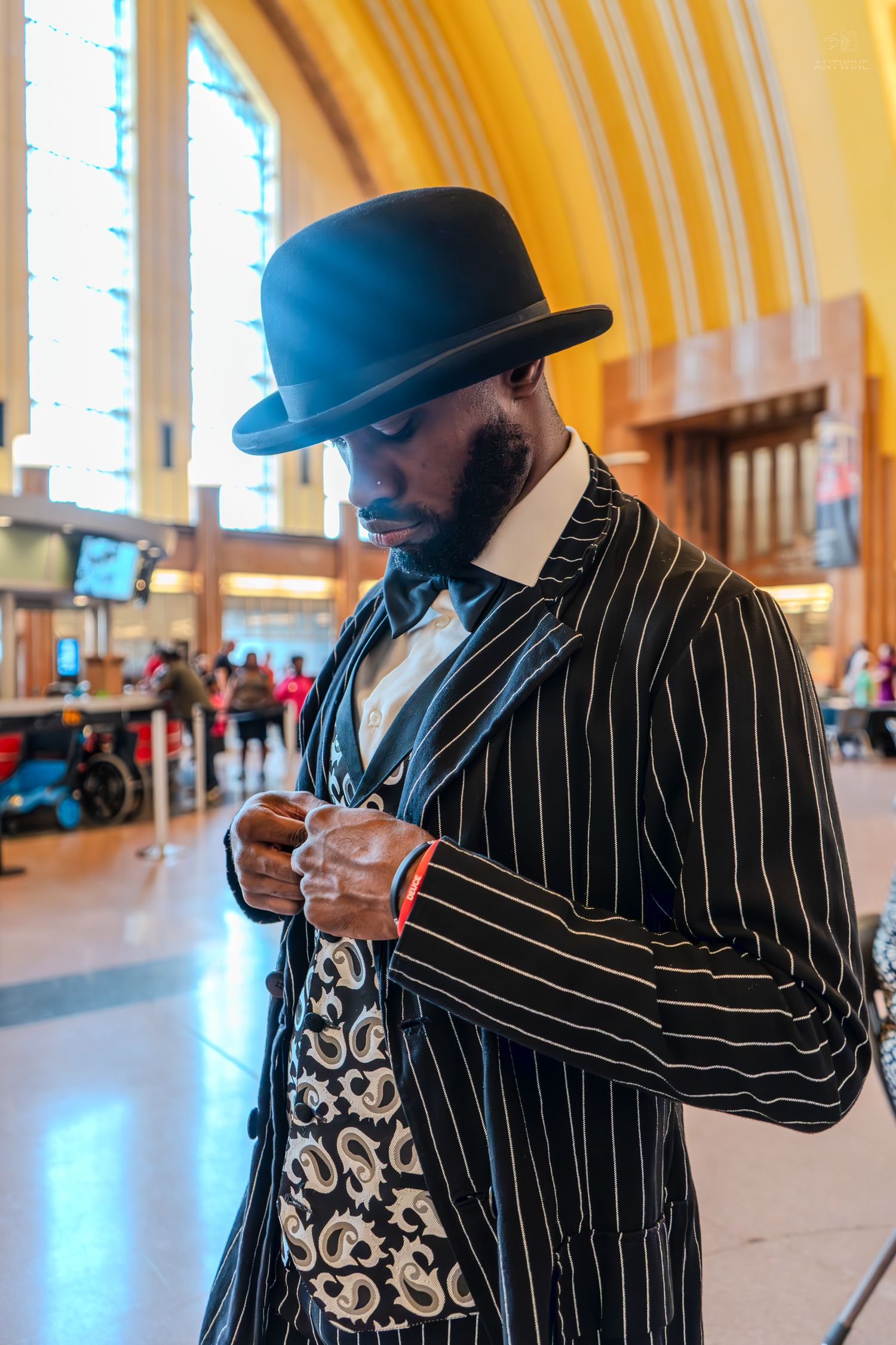 Dapper man in pinstripe suit and bowler hat adjusts cufflinks in ornate train station.