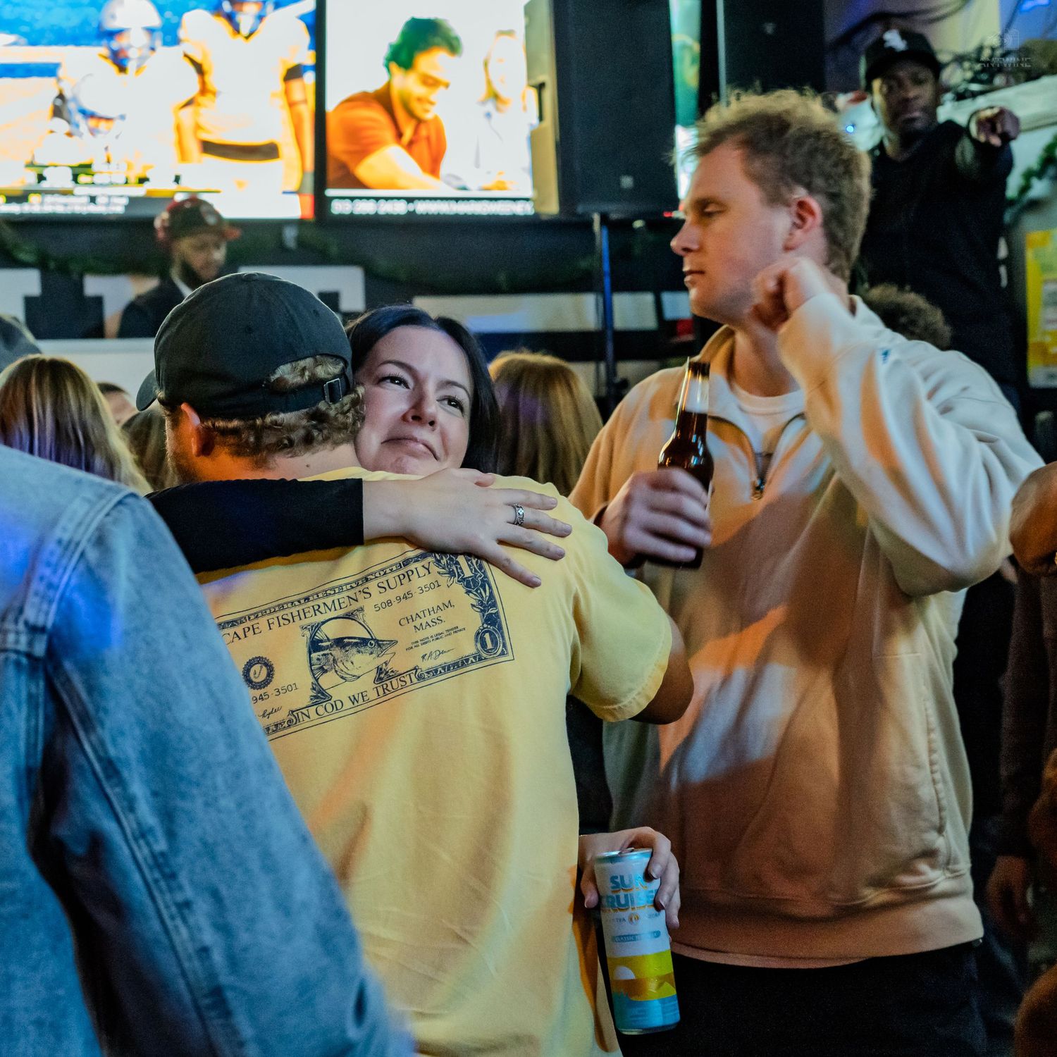 People hugging and socializing at a lively sports bar with large screens showing games in the background.