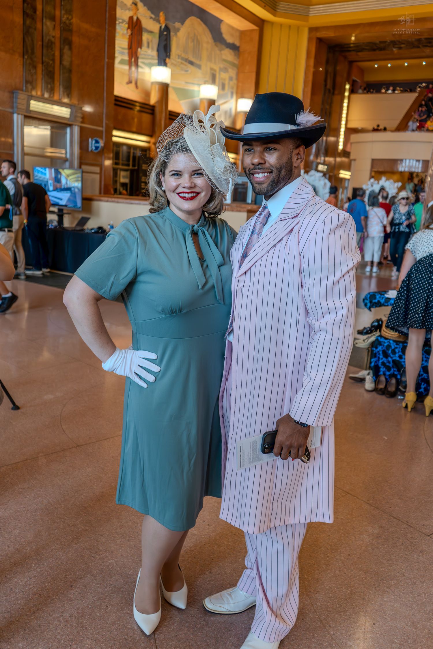 A couple in vintage 1940s attire pose indoors, woman in teal dress with fascinator, man in pinstripe zoot suit.