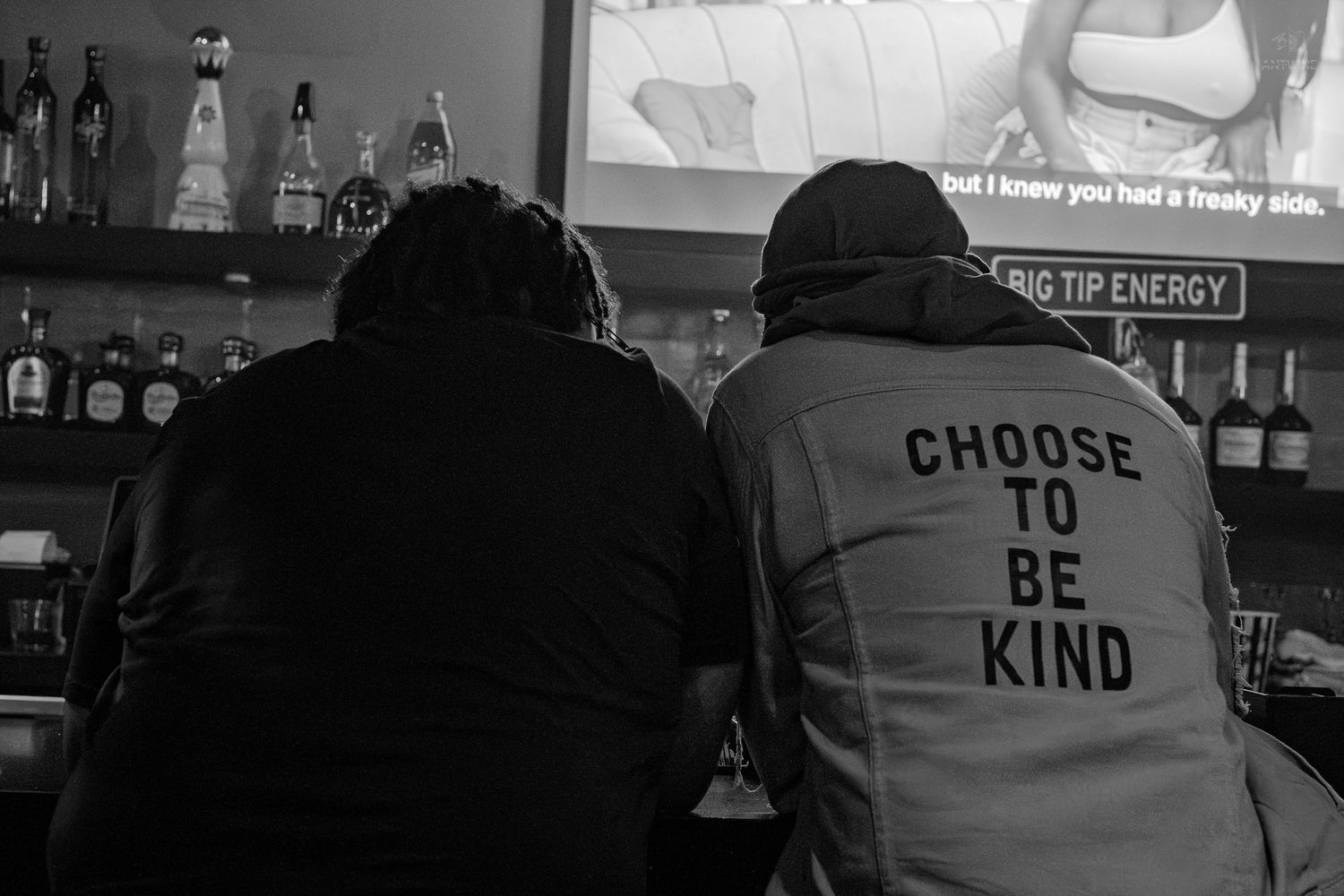 Two people sit at a bar, one wearing a hoodie reading Choose To Be Kind, in black and white.