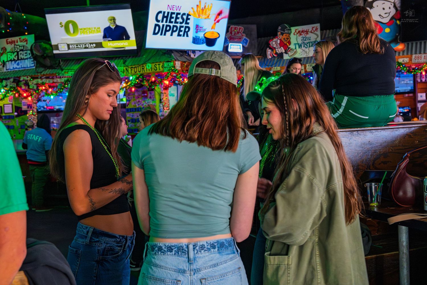 Three women socializing in a lively bar decorated with green lights and colorful signs.