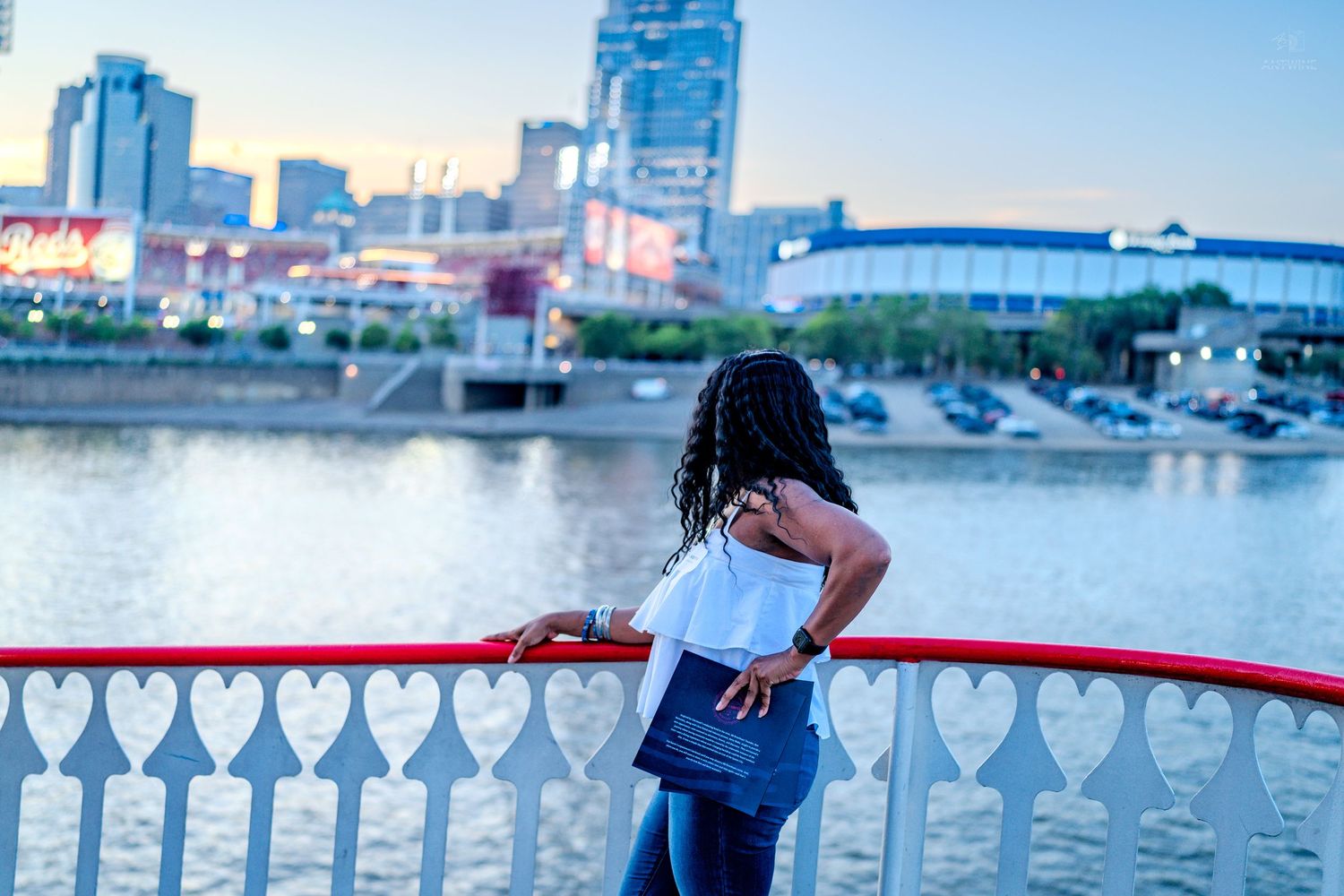 Woman leaning on red railing overlooking a river with city skyline at dusk.