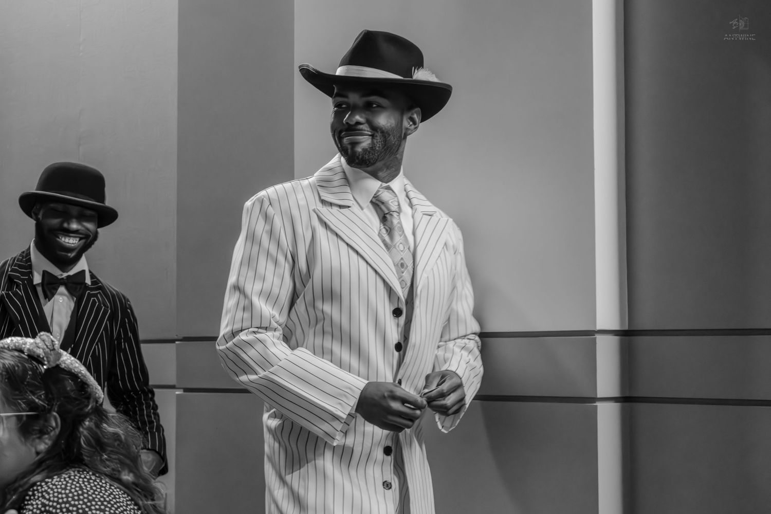 Man in pinstripe suit and fedora hat smiling at a formal event in black and white photo.