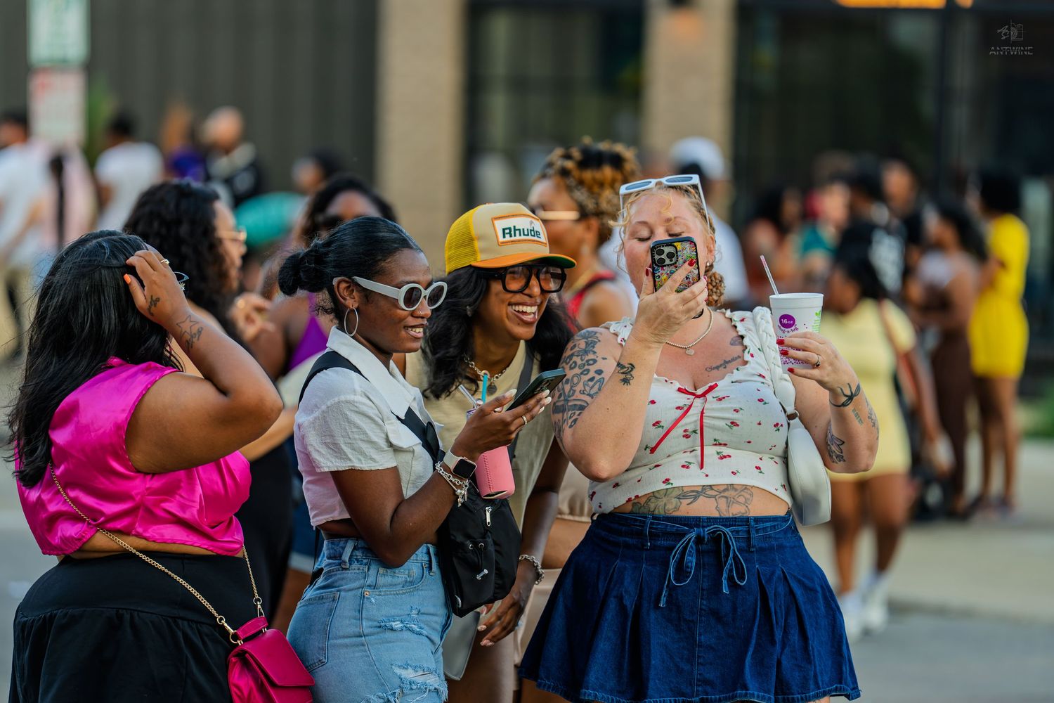 Women taking selfies and socializing outdoors while standing in a line at a summer event.