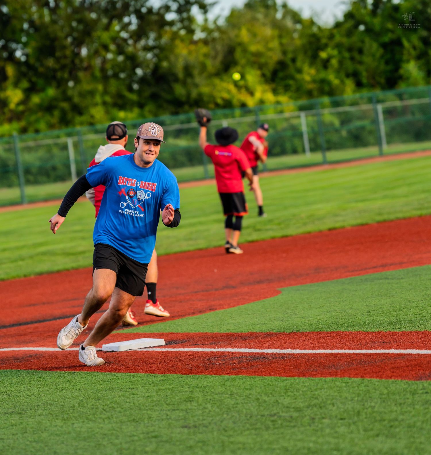 Softball player in blue shirt rounds third base smiling during an outdoor game on green turf field.