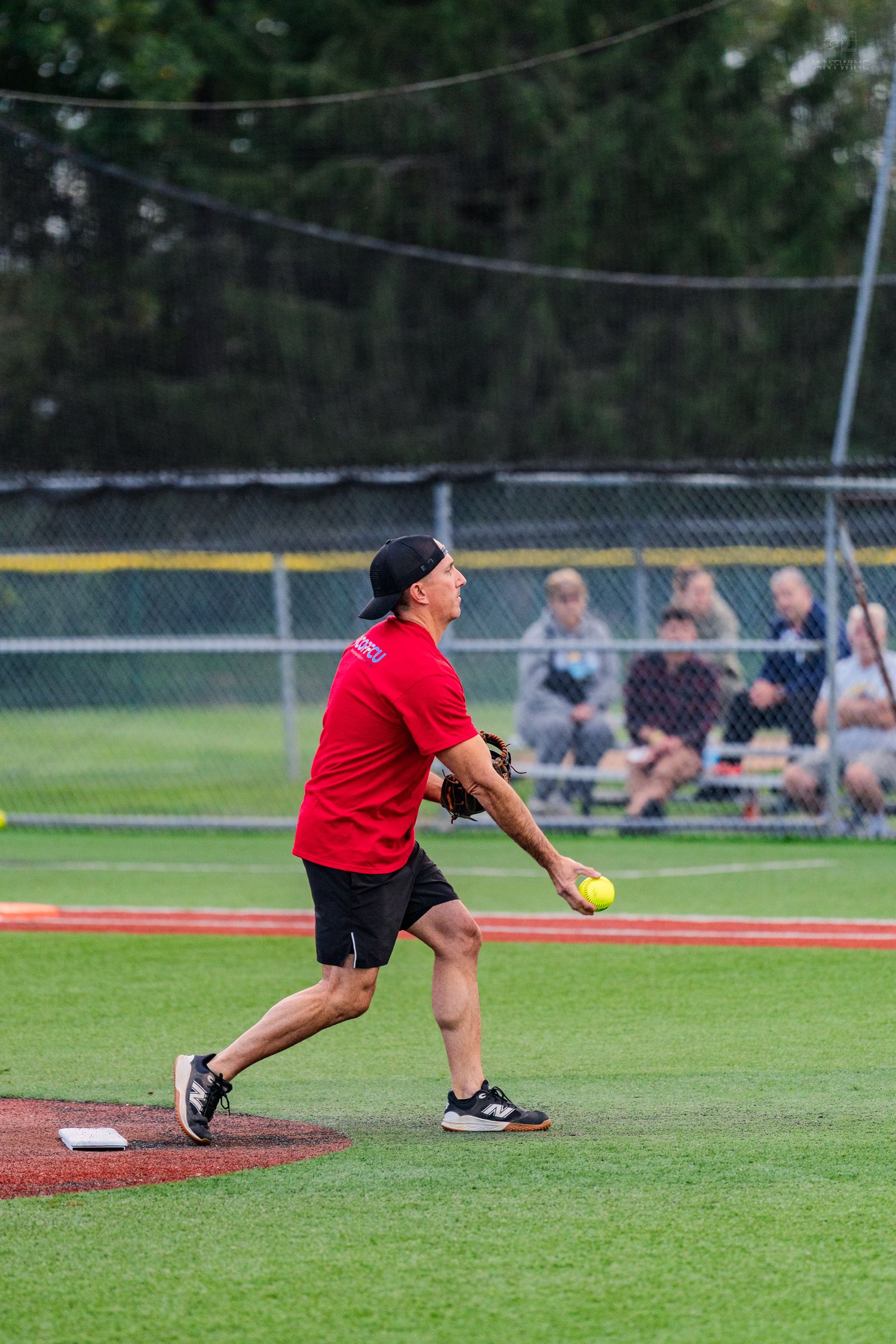 Softball pitcher in red shirt preparing to release yellow ball from pitching mound during game.