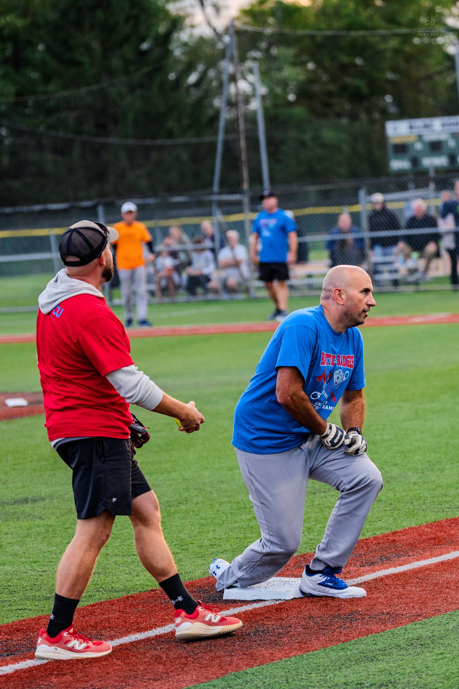 Two softball players in red and blue jerseys compete at first base during an evening recreational league game.