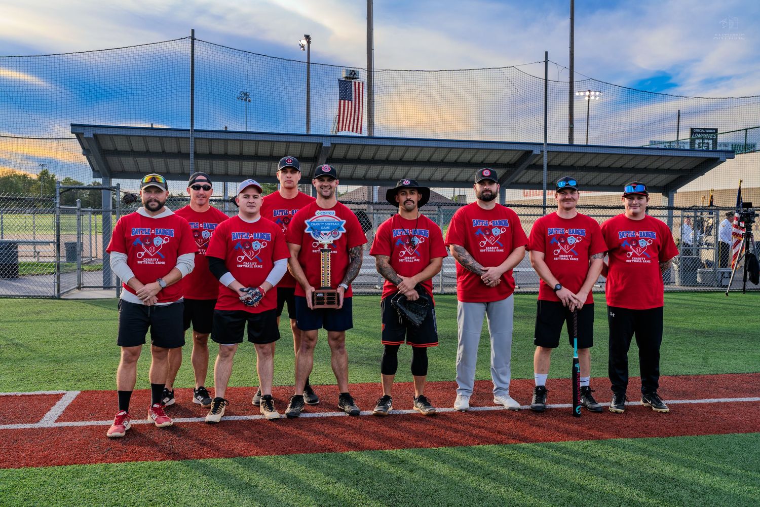 Youth baseball team poses with trophy on turf field wearing matching red championship shirts at dusk.